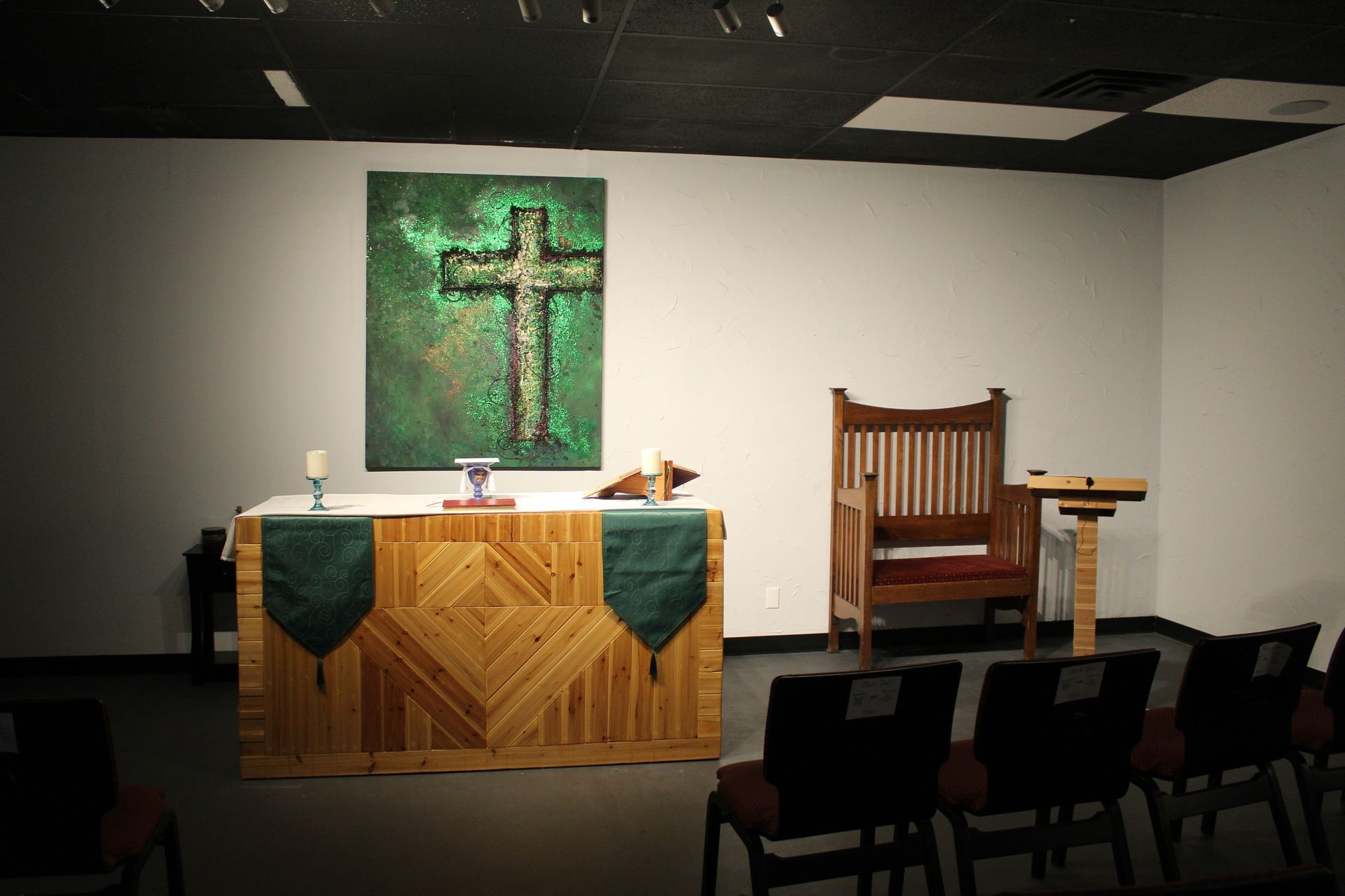 A church altar with cross painting, wooden table with green cloth, chair, and podium.