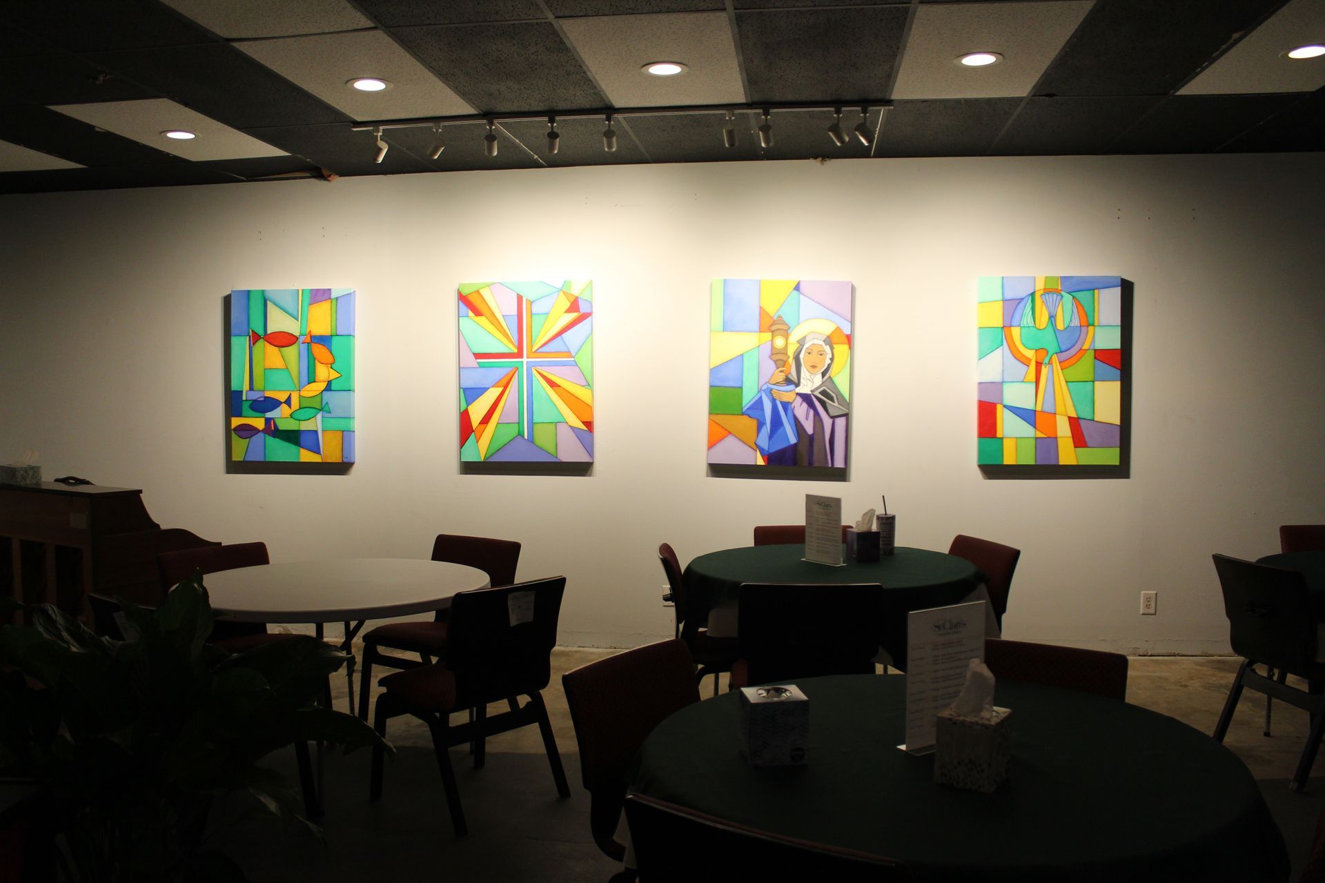 Gallery with four colorful paintings on a white wall, tables, chairs, and recessed lighting.