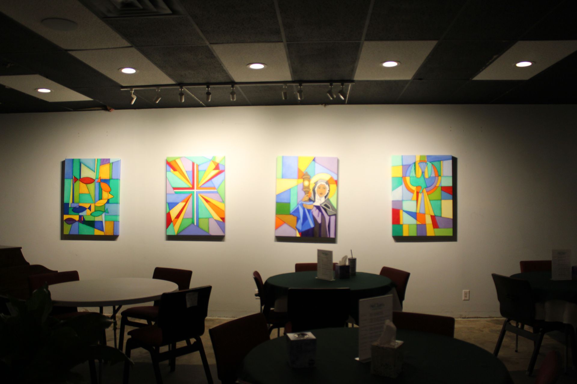 Four colorful abstract paintings hang on a wall above round tables and chairs in a dimly lit room.