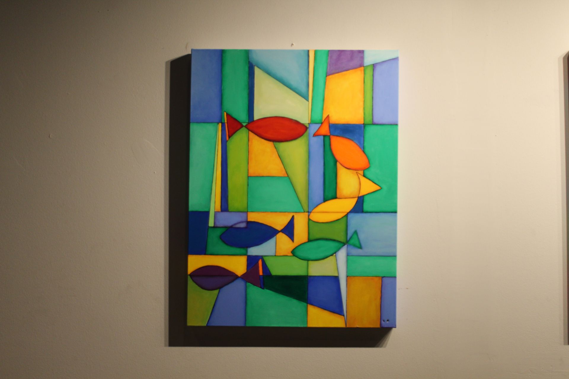 Abstract painting of fish. Colorful, geometric shapes in greens, blues, orange, and yellow. Hung on a wall.