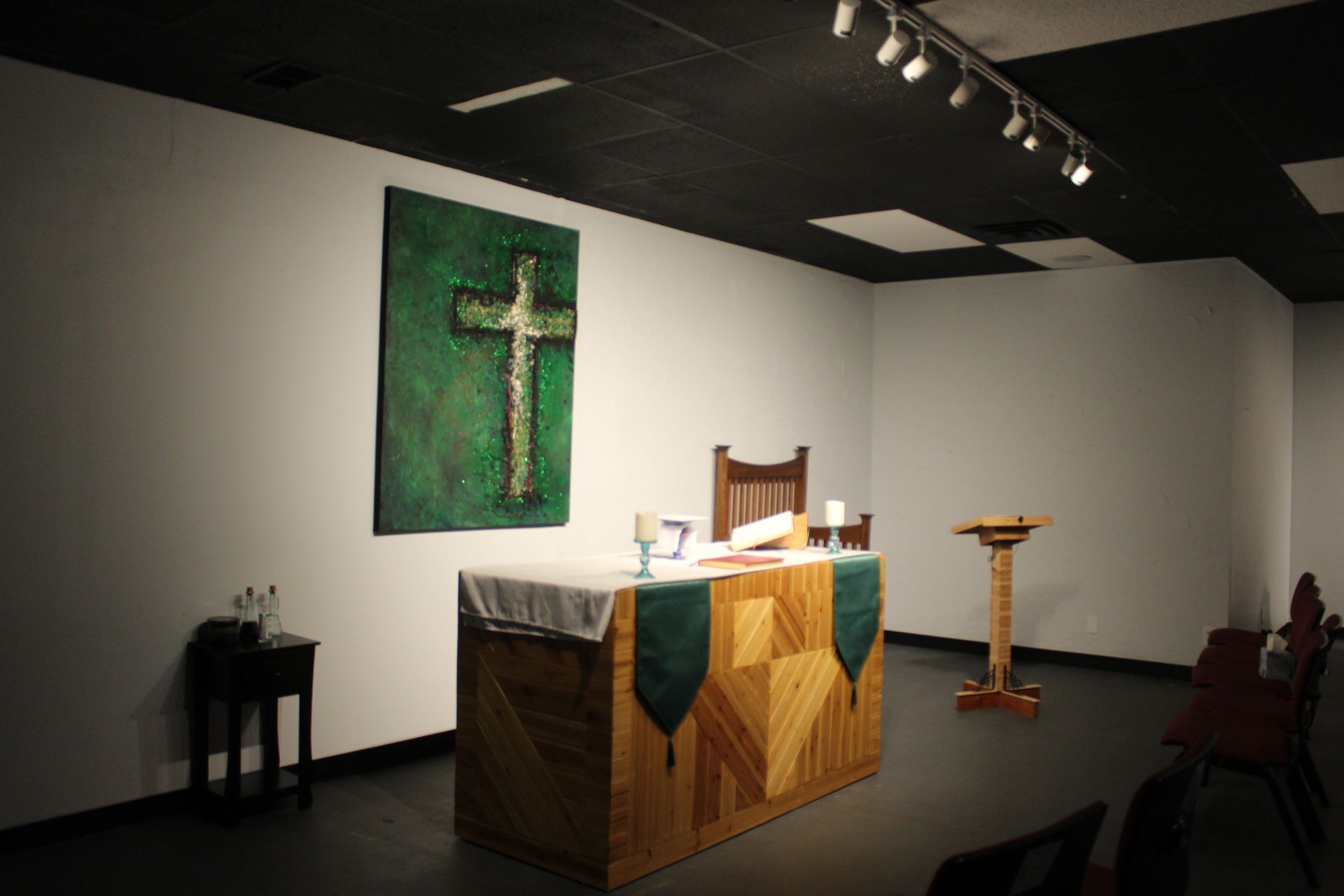 A dimly lit chapel with a wooden altar, green cross art, and a lectern; chairs along the right wall.