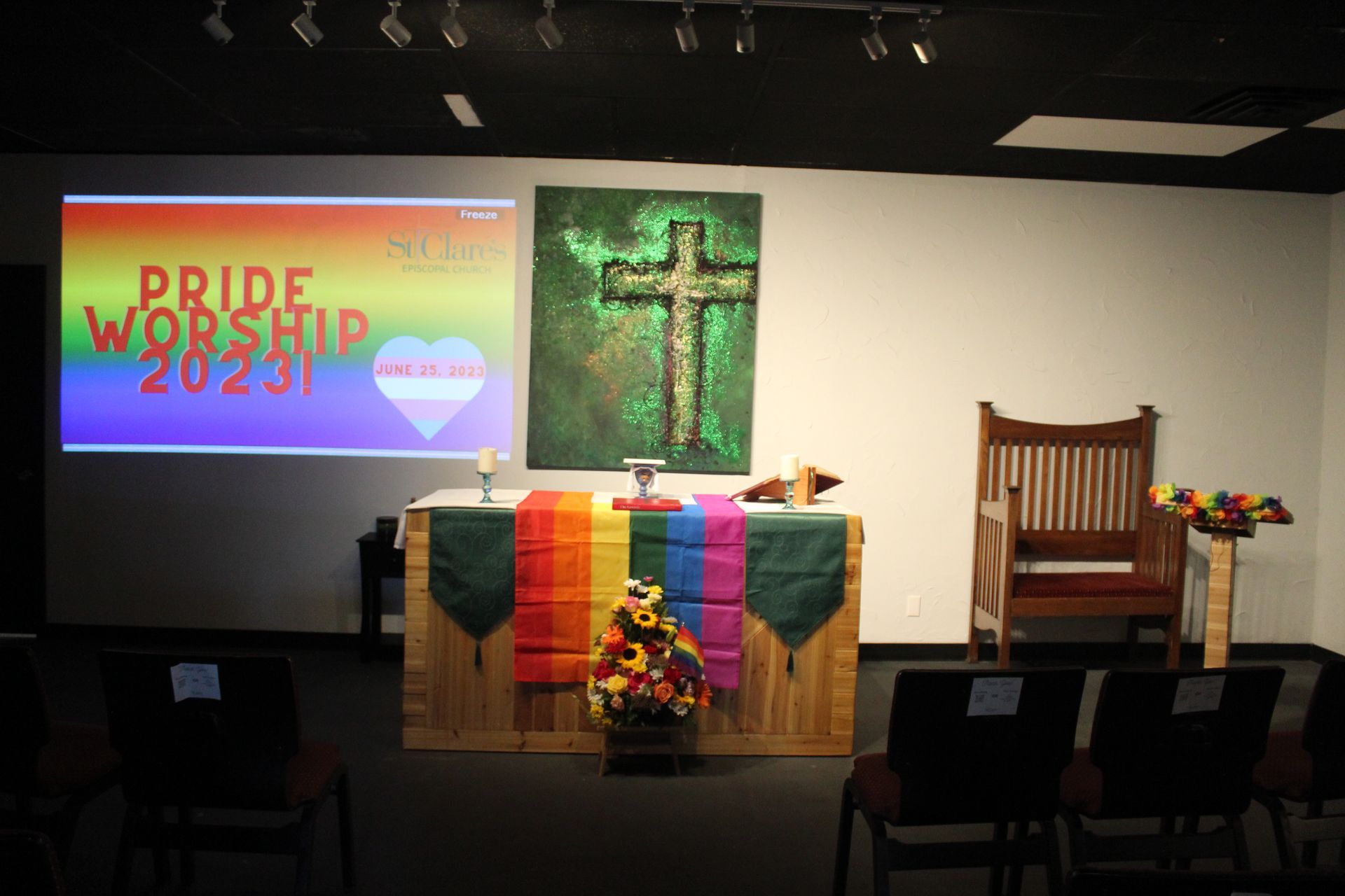 An altar with rainbow-colored stole, a Pride Worship 2023 presentation, a green cross, and chairs set up in a room.