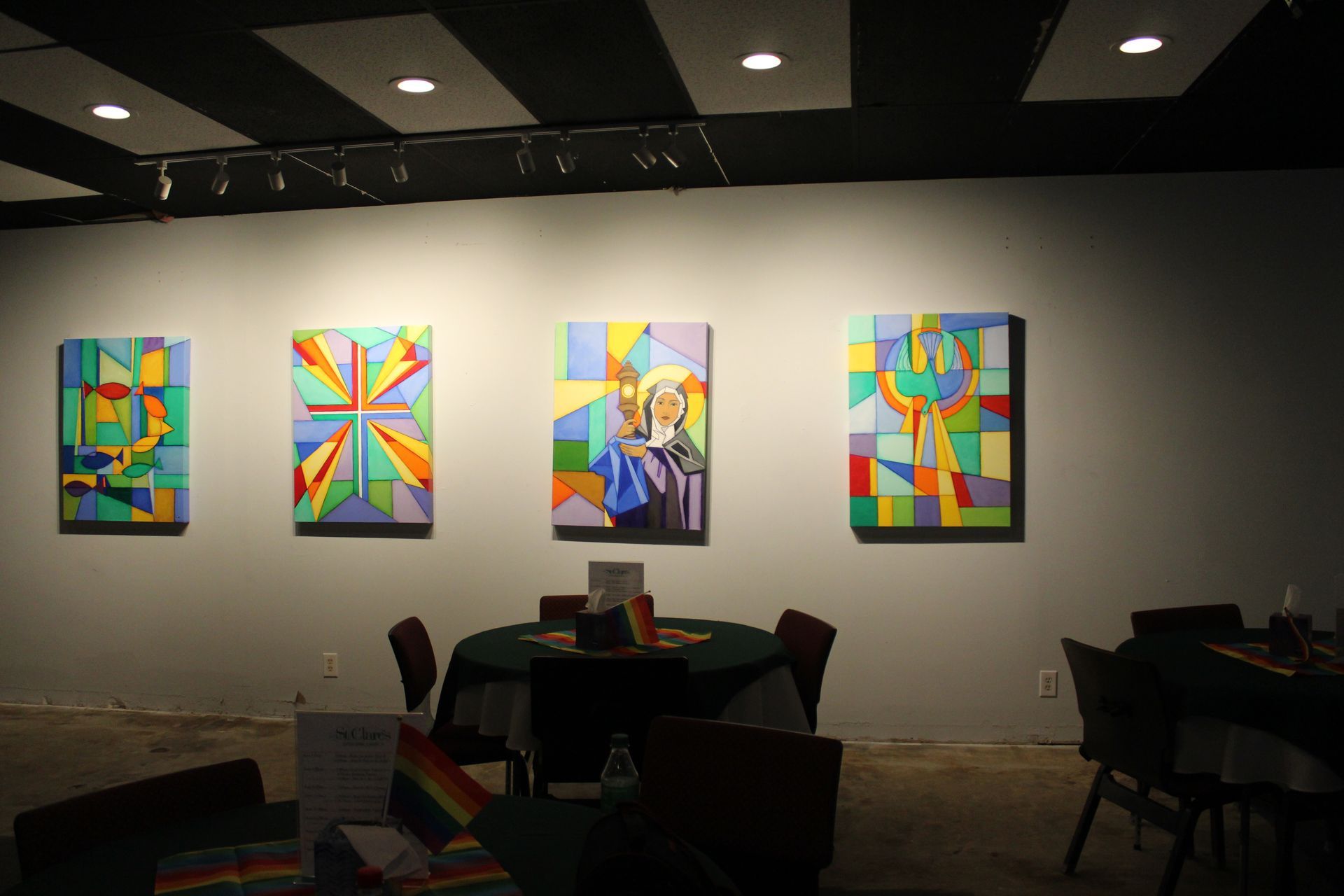 Four colorful abstract paintings on a gallery wall, a table with a rainbow cloth.