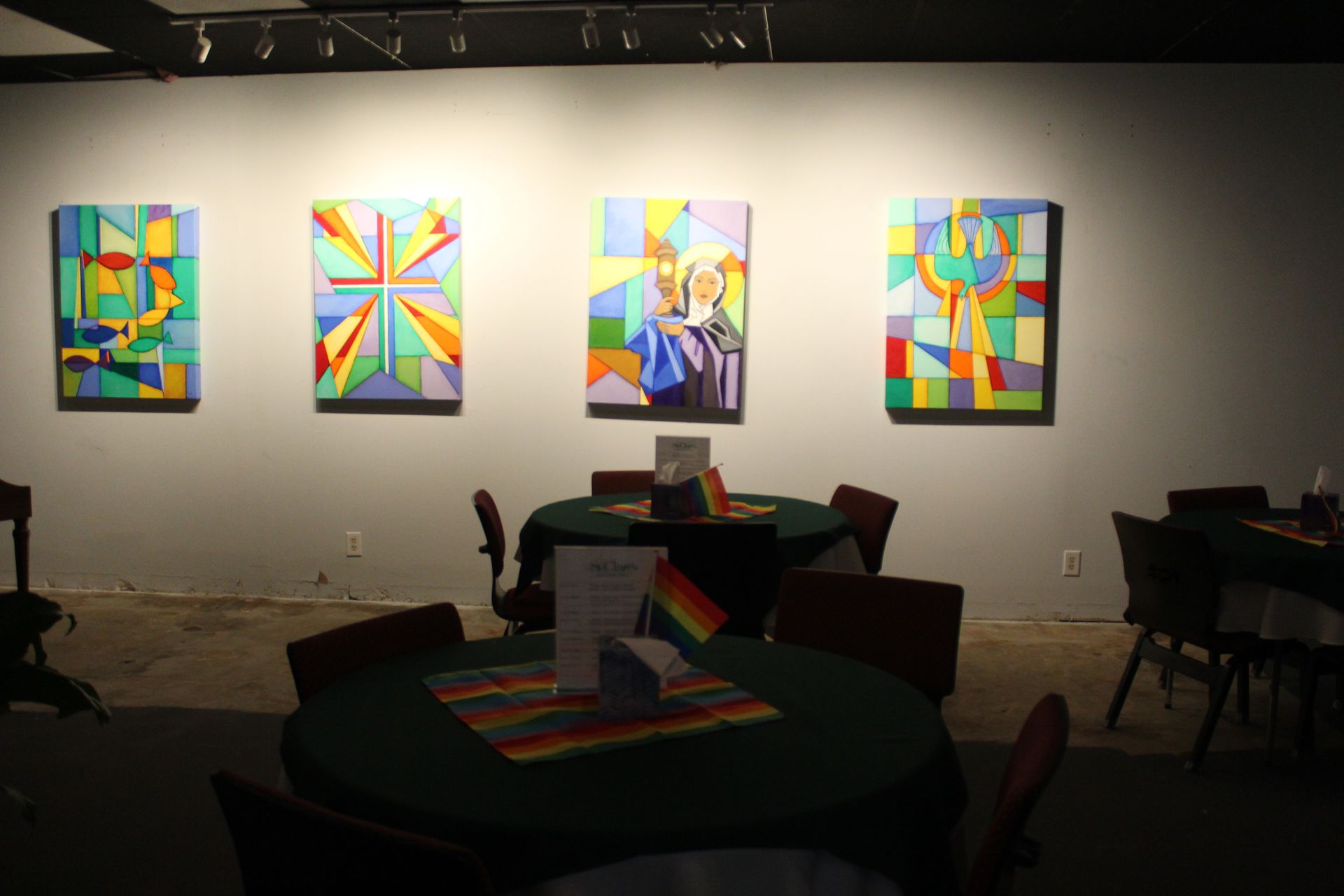 Gallery with four colorful paintings on the wall; round tables with green cloths and chairs in the foreground.