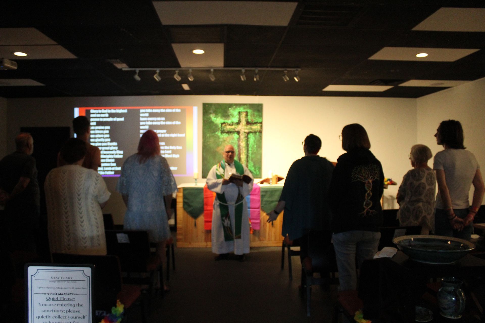 A religious service with a pastor at an altar. People stand, looking toward the cross and presentation screen.