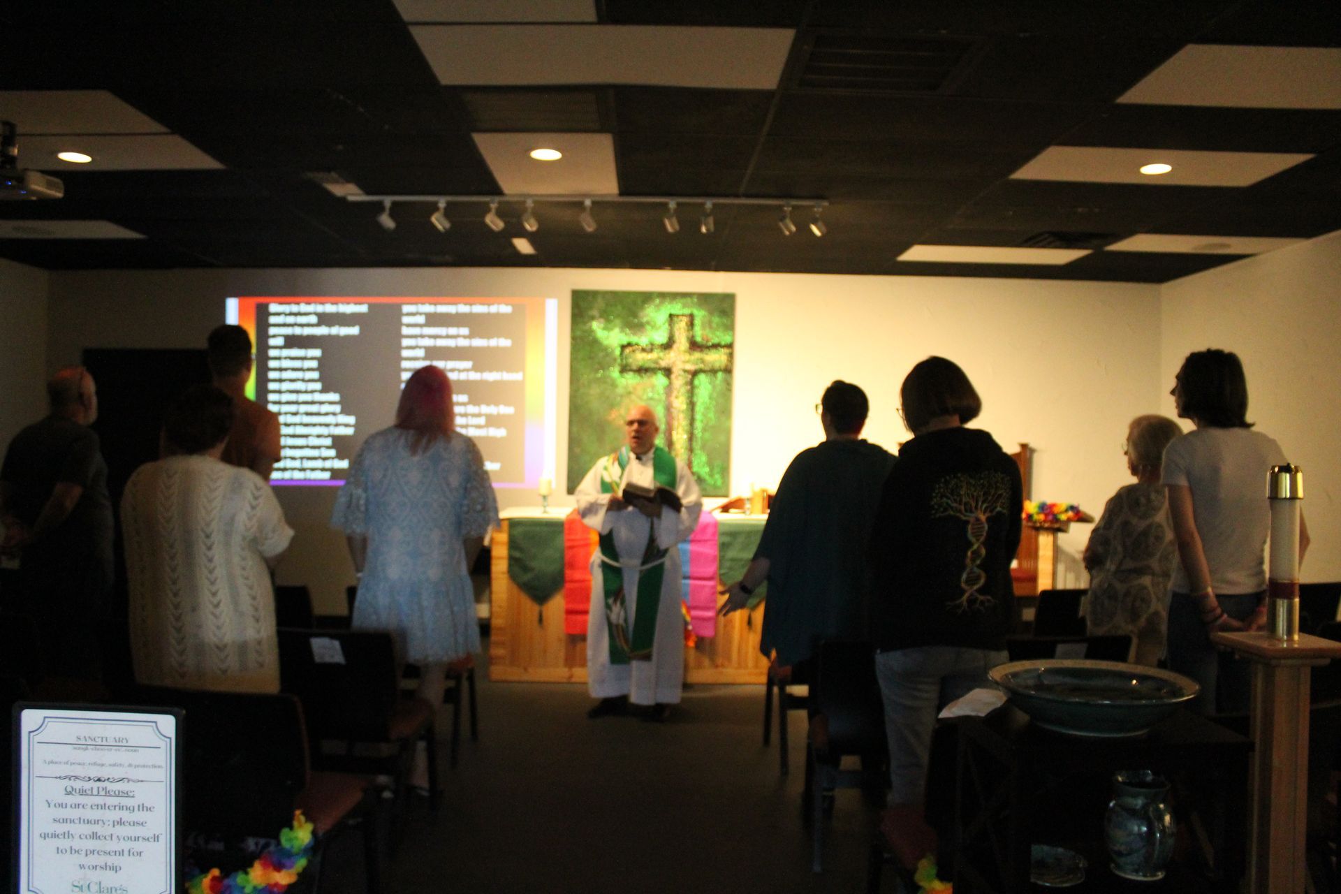 People standing and facing a priest at a church altar. A cross hangs on a green tapestry. Projection screen visible.