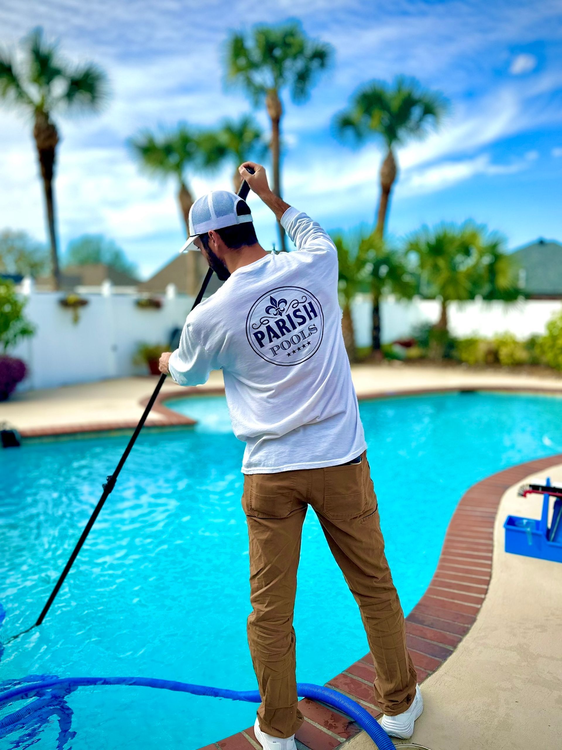 Man cleaning a pool with a long-handled brush, wearing white shirt, tan pants, and a white hat.