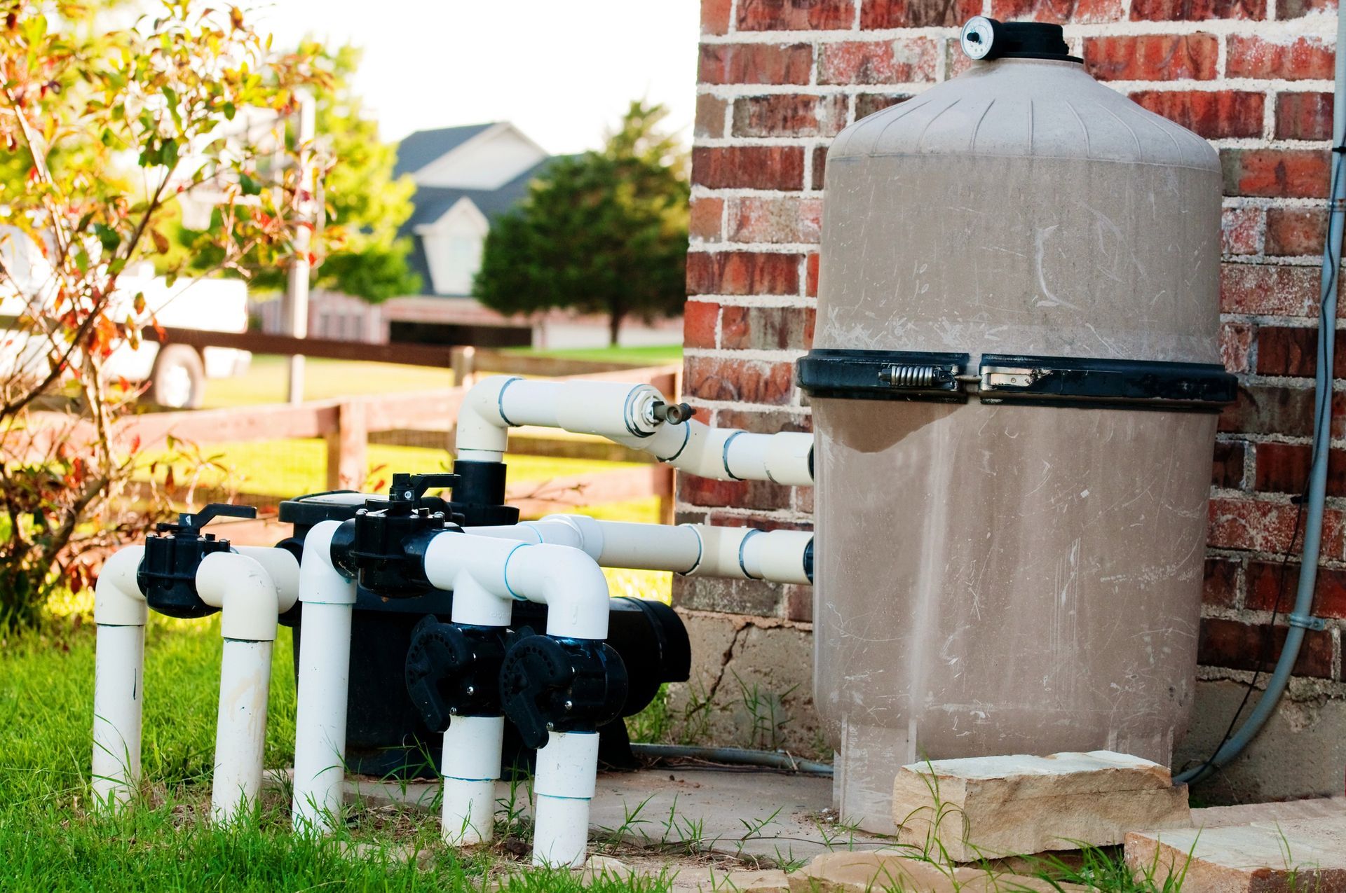 Pool filter system with pipes and a tank, set against a brick wall and grassy yard.
