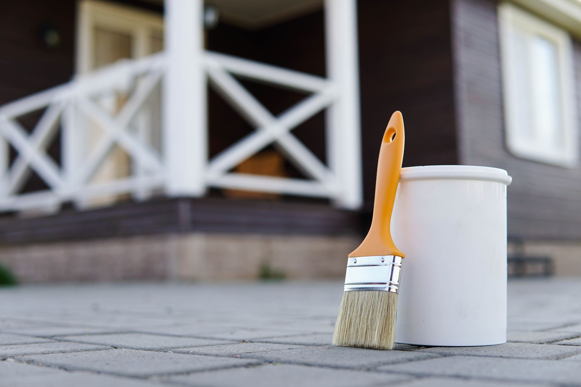 A brush and a can of paint are sitting on the sidewalk in front of a house.