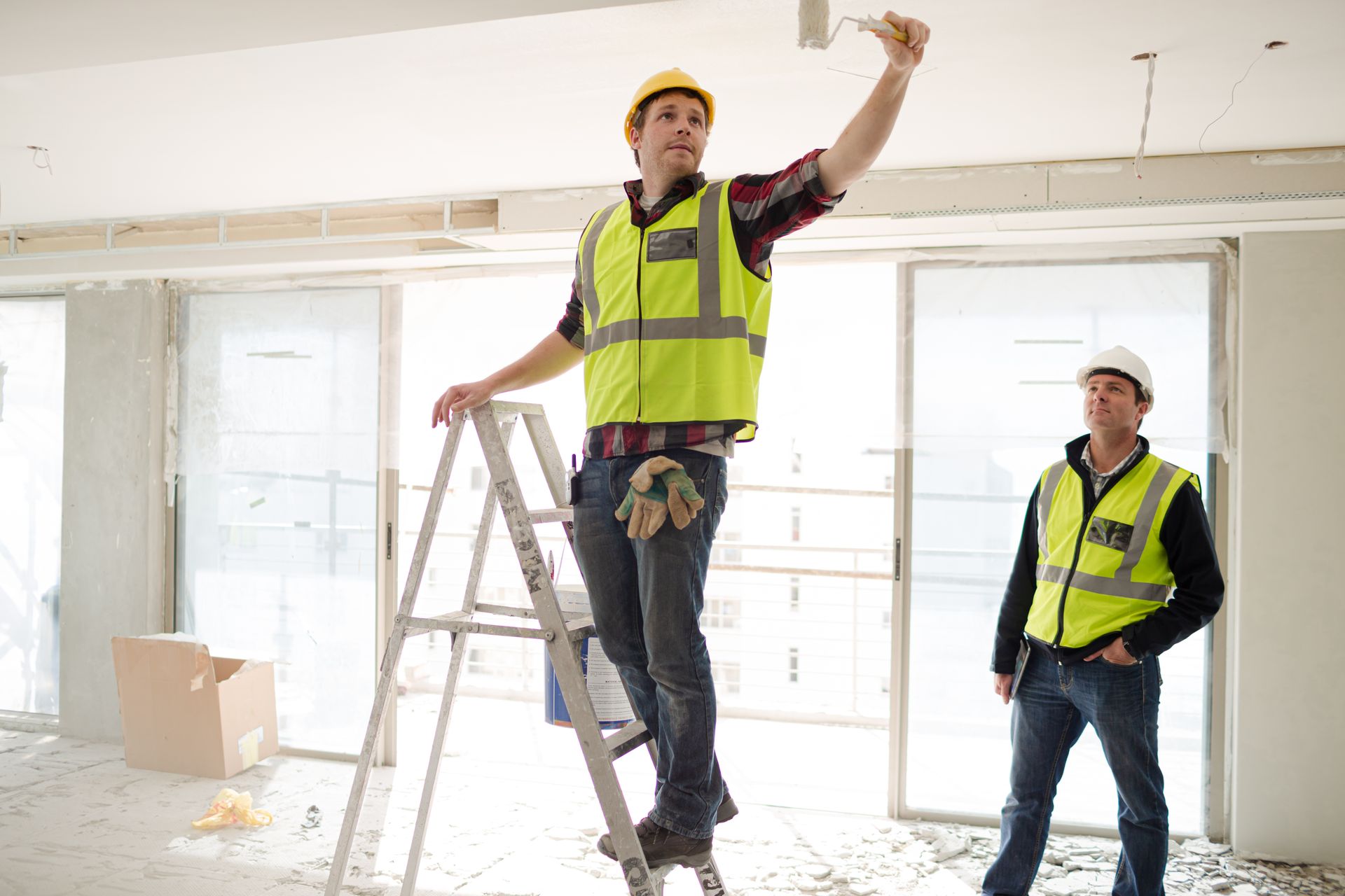 Two construction workers painting a ceiling; one on a ladder, the other standing near; both wearing safety vests and helmets.