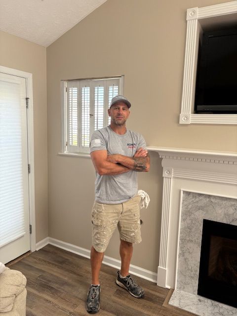 Man in grey shirt, khaki shorts, and baseball cap stands in front of a fireplace with arms crossed.