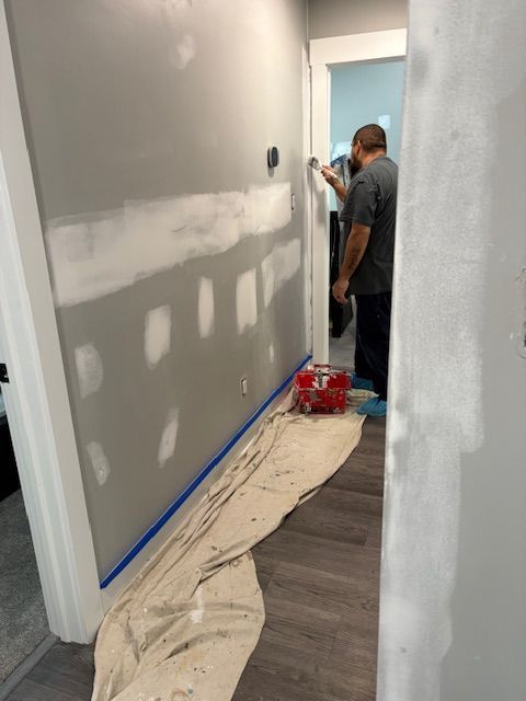 Man painting a gray wall in a hallway, covering patches of white spackle. Blue tape and drop cloth protect the floor.