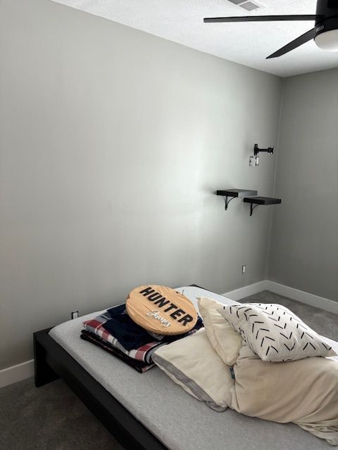 Bedroom with bed, pillows, blanket, shelves, and a ceiling fan. Gray walls and carpet.