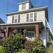 Two-story house with gray siding, red shutters, and a porch. American flag and bushes in front.
