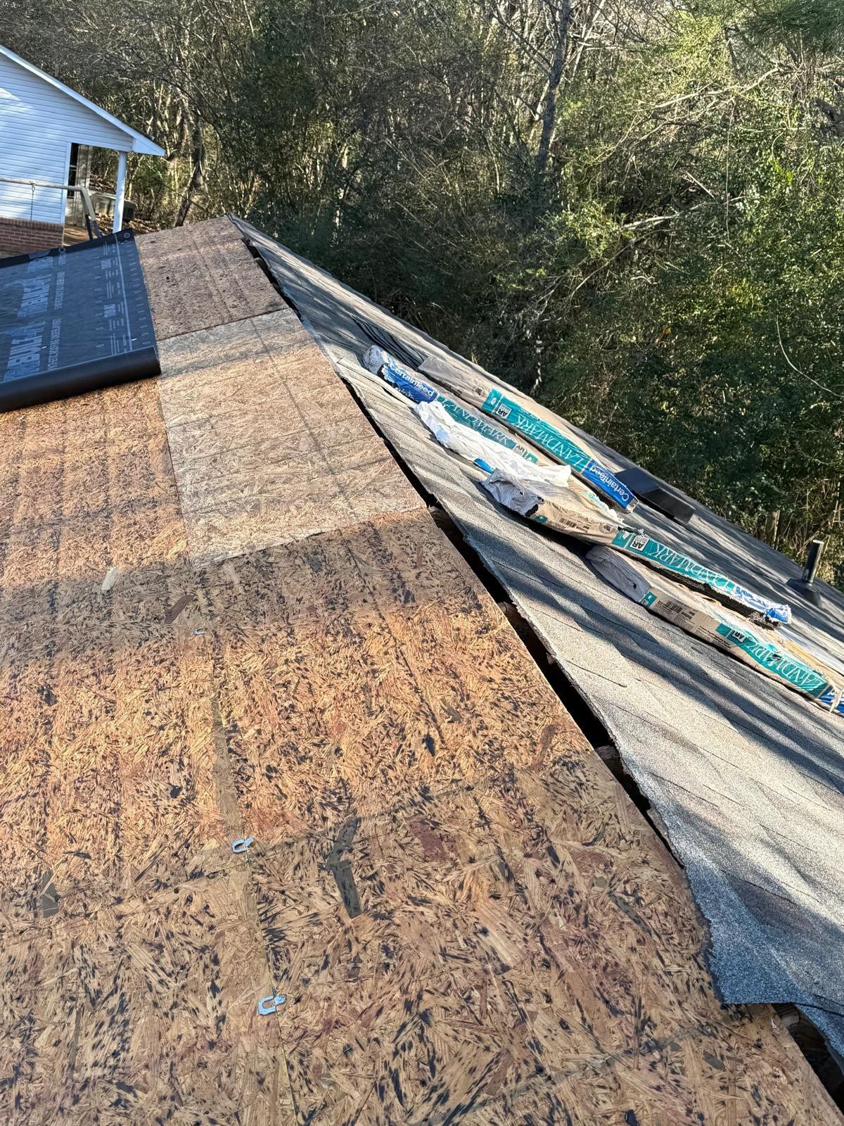 Roof with exposed wood and damaged shingles; green trees in background.