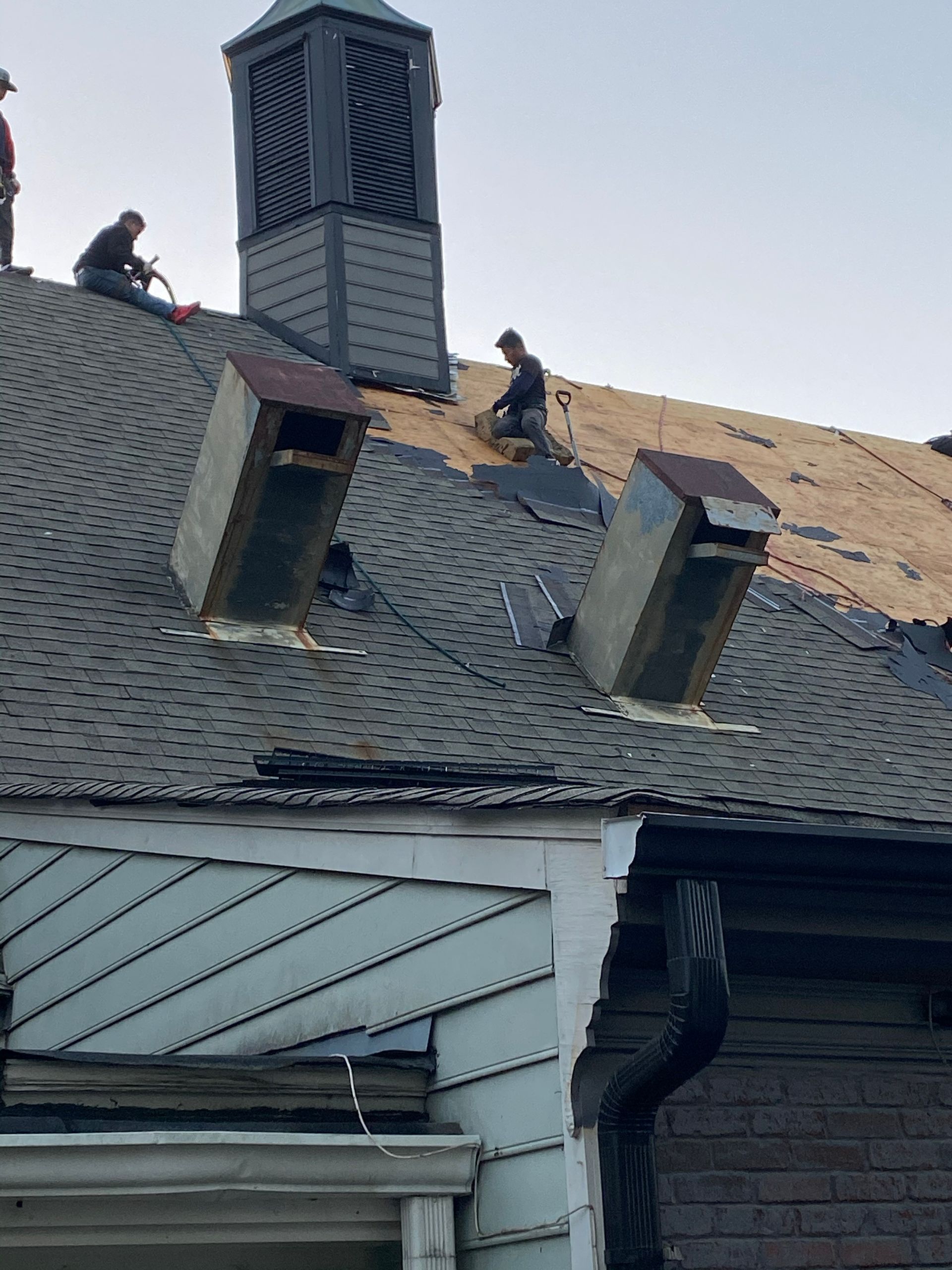 Roofers working on a house roof, removing old shingles and installing new ones; gray sky.