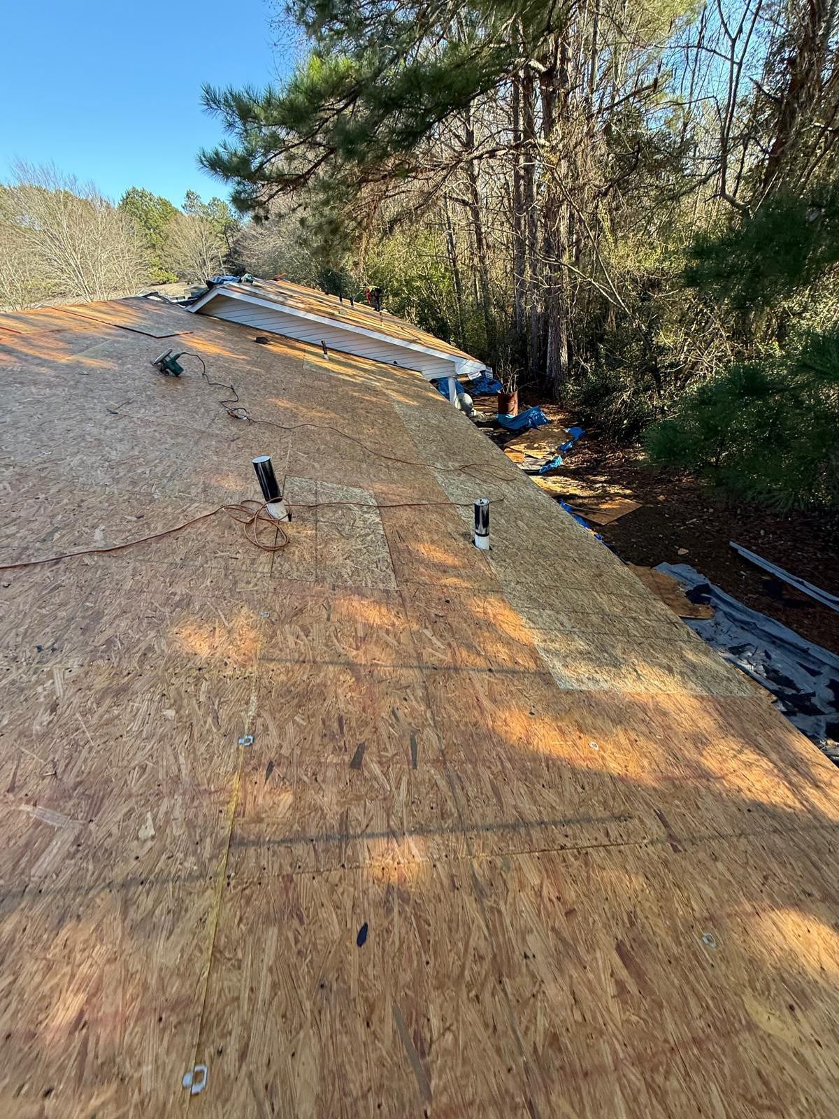 Rooftop with exposed wooden sheathing and metal flashing, trees in the background, sunny day.
