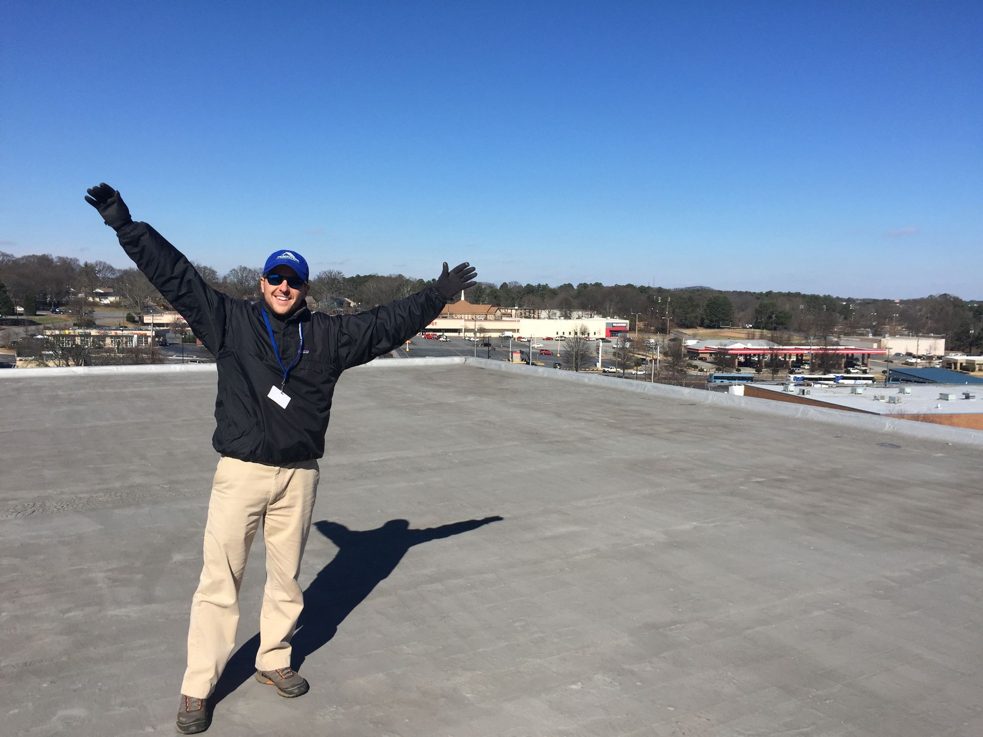 Man on rooftop with arms raised, sunny day. Gray roof, blue sky, buildings in background.