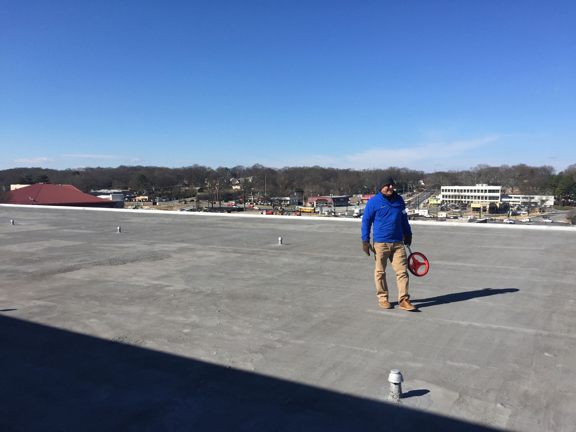 Person in blue jacket on a flat roof, holding a measuring wheel, with town in background on a sunny day.
