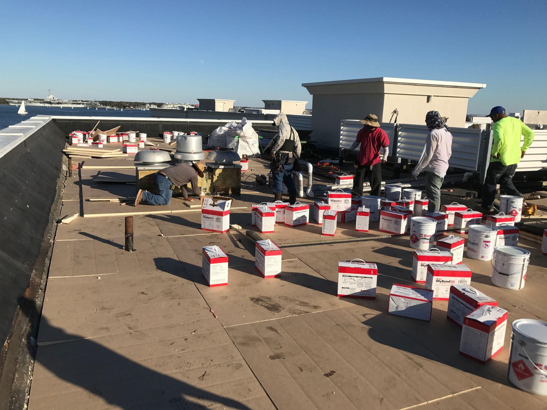 Construction workers on a flat roof with materials, cans, and a harbor view.