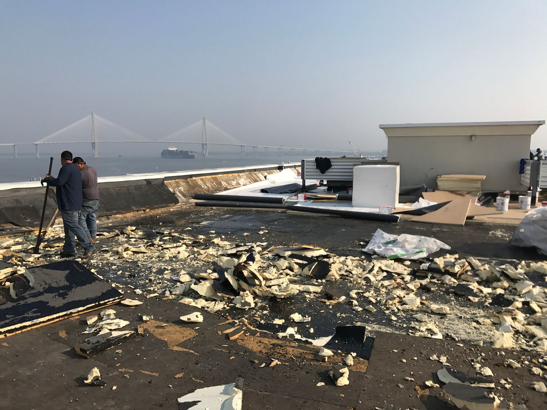 Two people on a rooftop with debris, possibly construction. A bridge and water are in the background.