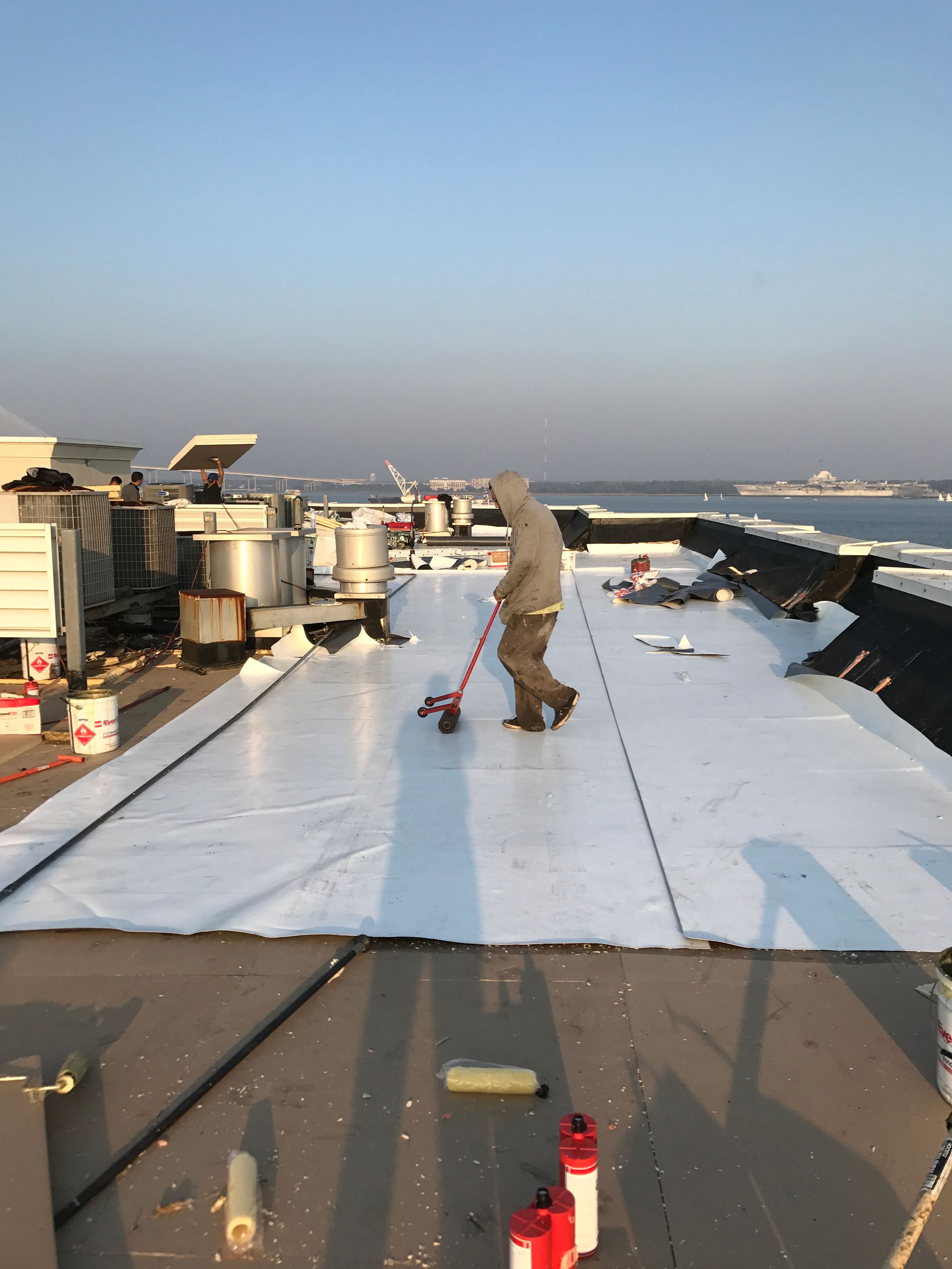 Person on a roof using a tool to smooth out white roofing material. Daytime setting with other rooftop equipment.