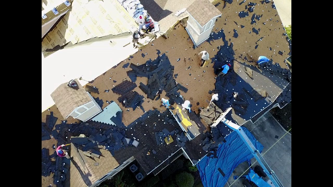 Aerial view of workers on a roof, tarp and debris visible.