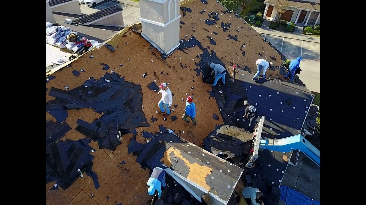 Roofers working on a residential rooftop. Brown shingles, white chimney, blue tarps, and workers with safety hats.