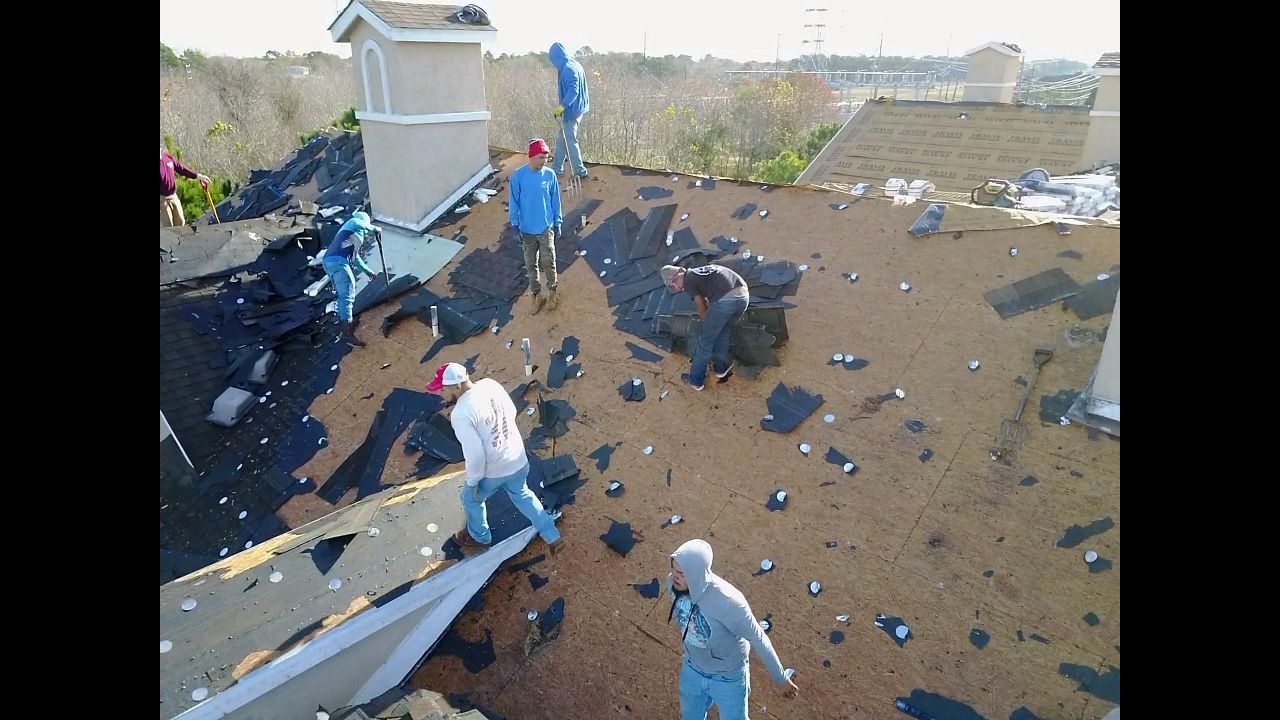 Roofers removing shingles from a rooftop. Several people working in daylight, debris scattered around the roof.