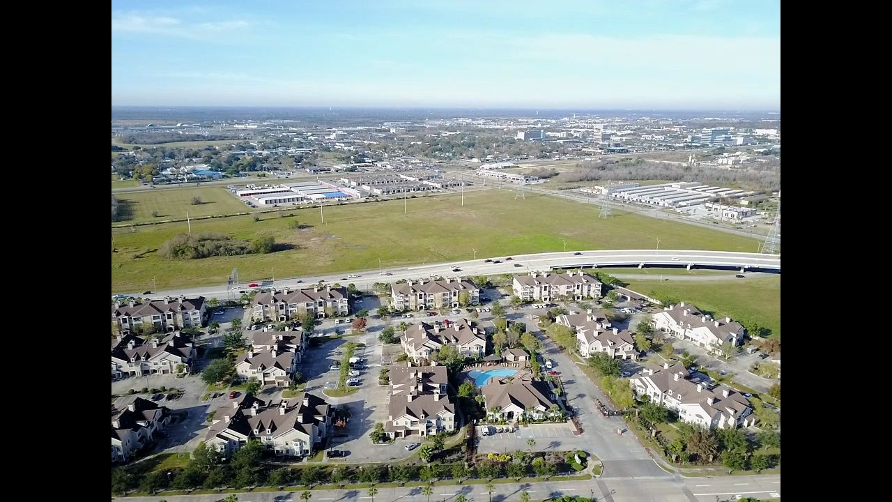 Aerial view of a suburban neighborhood with tan buildings, pool, and highway, with a city in the distance under a blue sky.