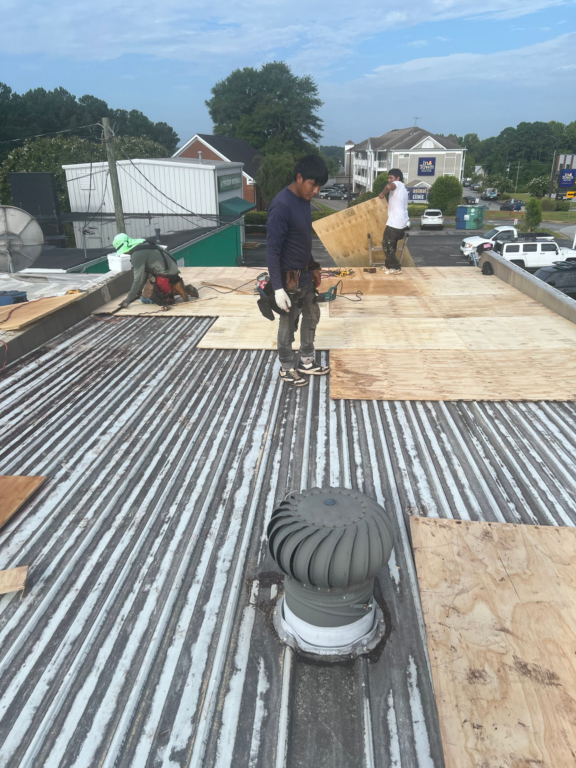 Workers replacing plywood on a corrugated metal roof; one man holds wood; a roof vent is in the foreground.