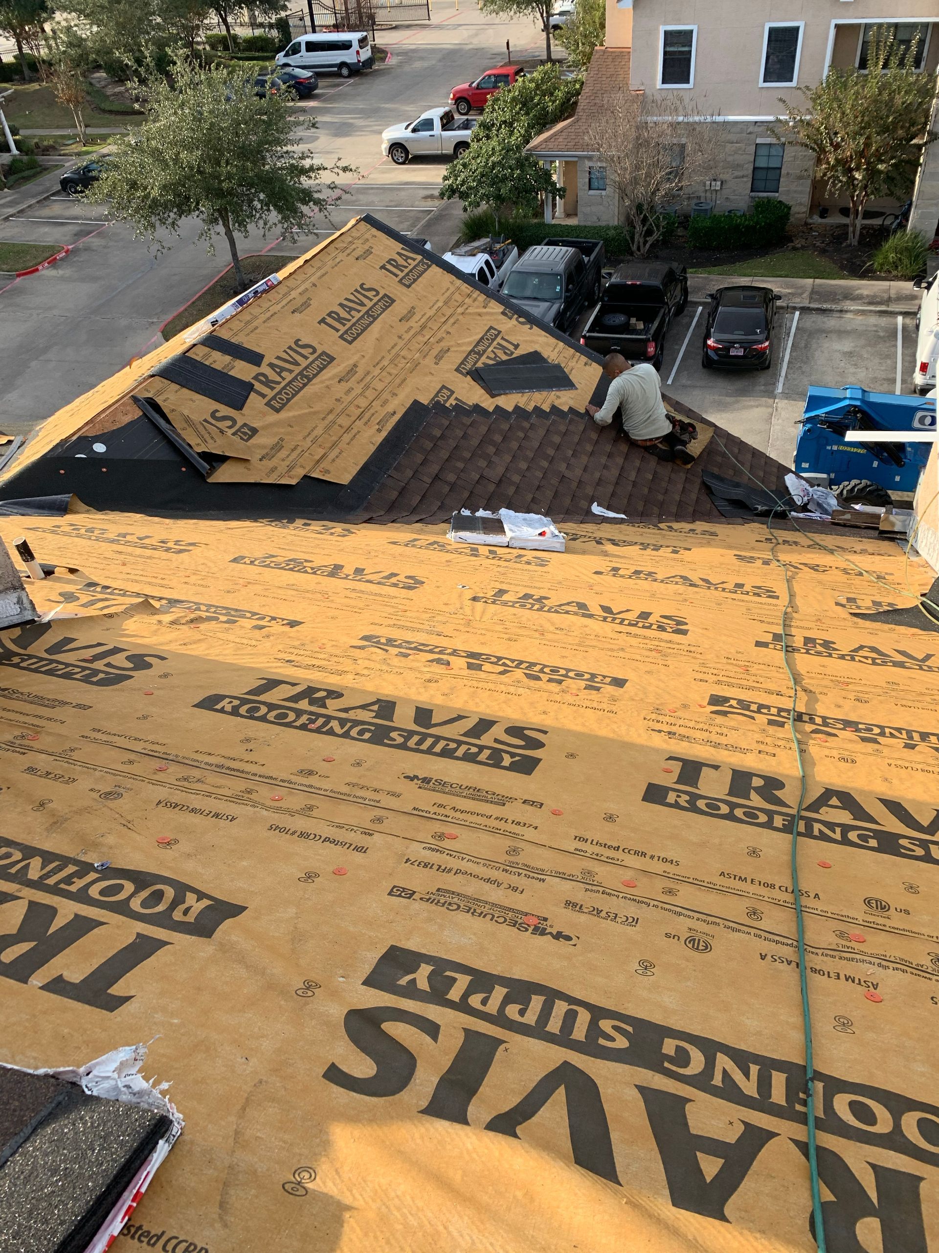 Roofer on a residential roof, installing shingles. Brown roofing felt and asphalt shingles. Cars parked below.