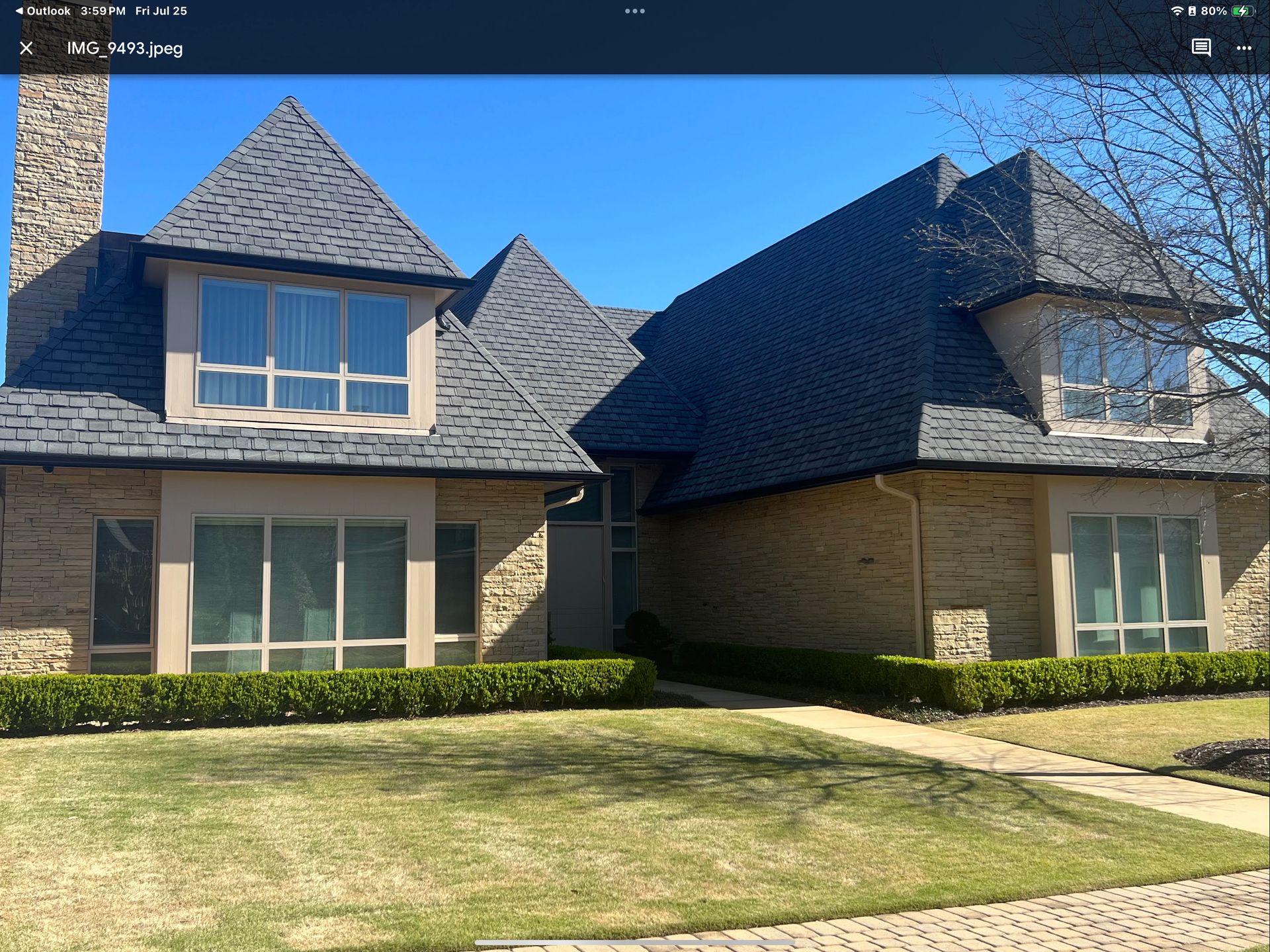 Tan brick house with dark shingle roof, green lawn, clear sky, and large windows.