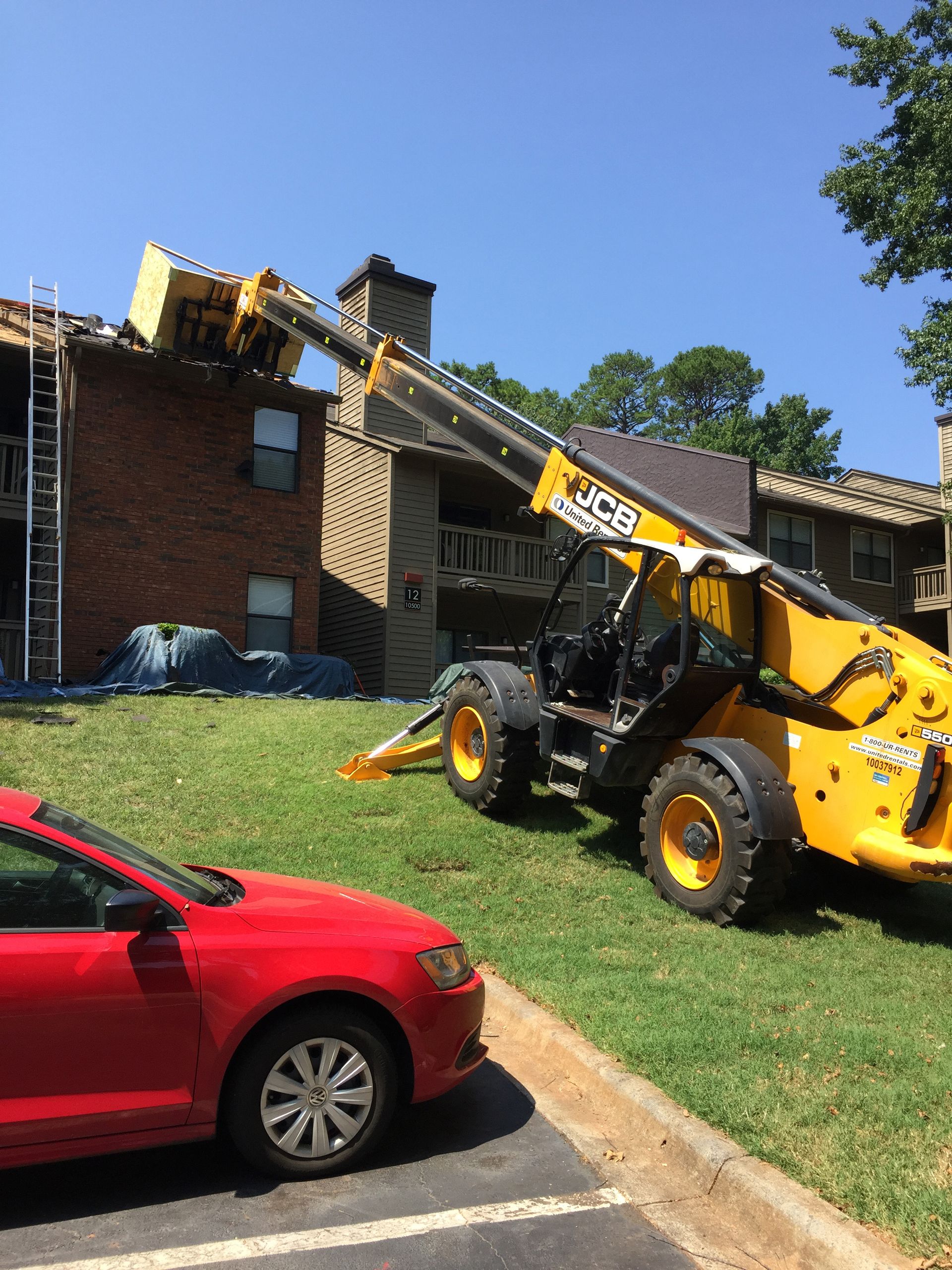 Yellow telescopic forklift removing something from a building's roof; a red car is parked nearby.