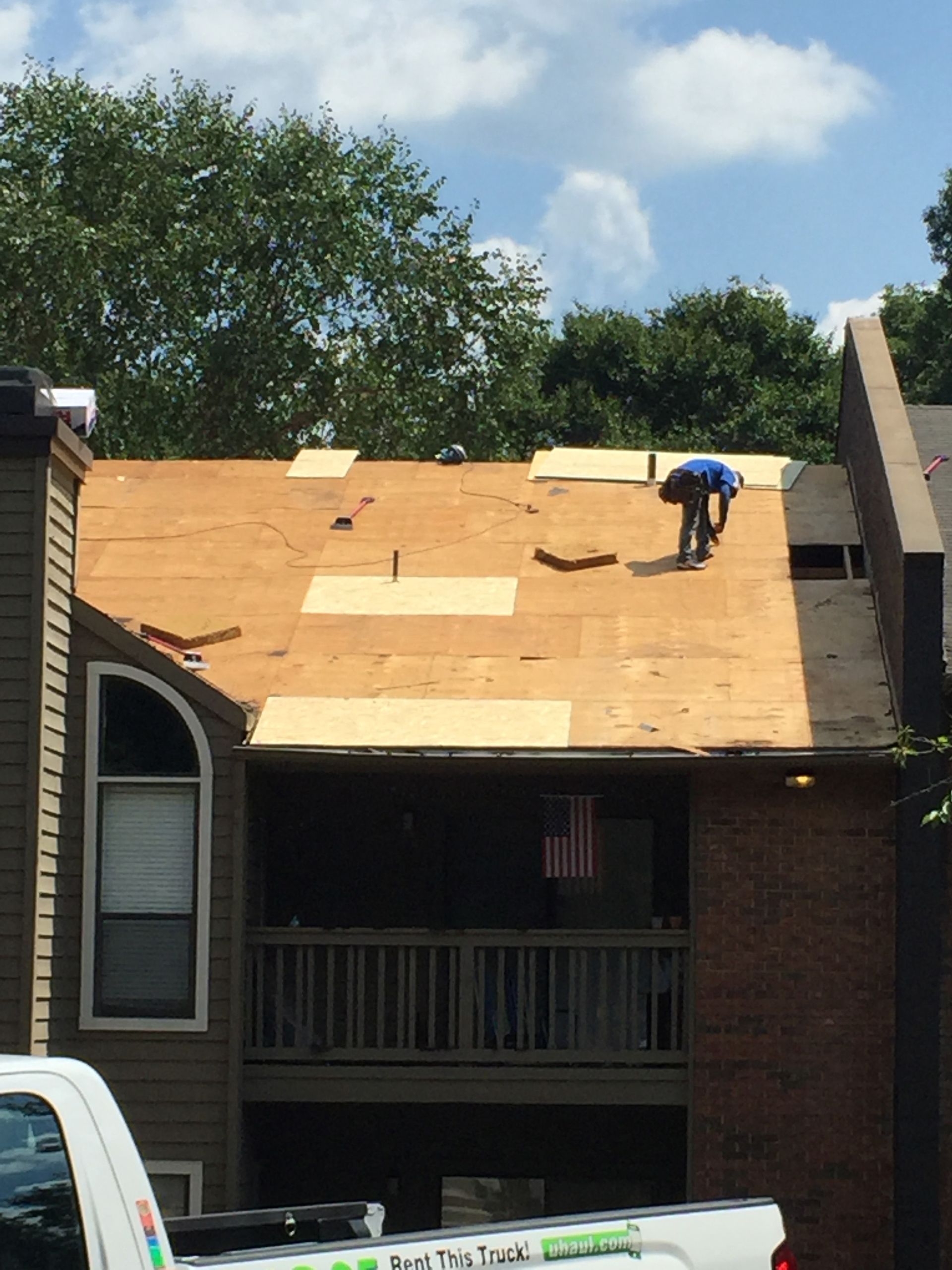 Roofer working on a partially shingled roof. Beige roof, blue sky, and green trees.