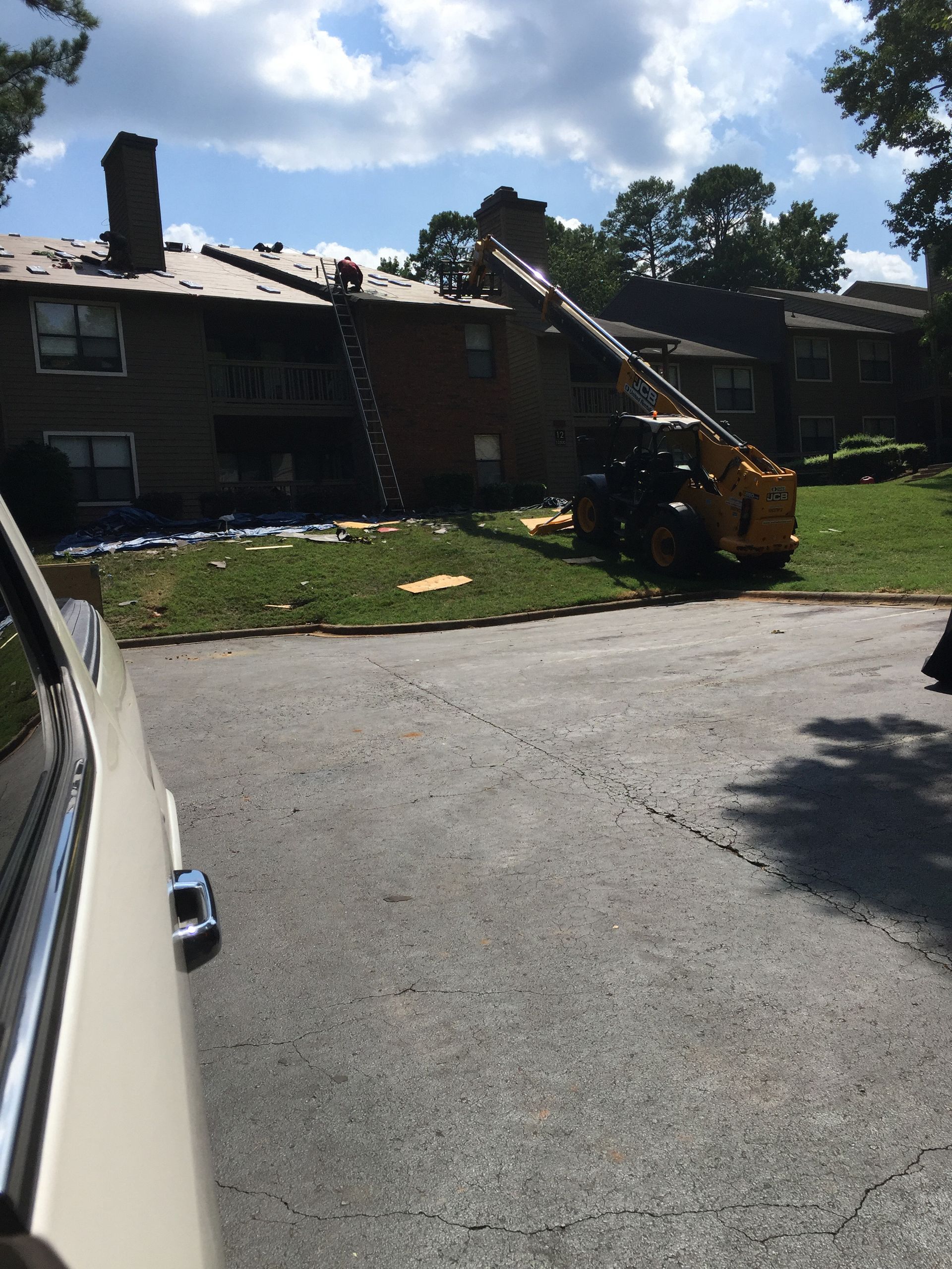 Crane and workers on a roof, possibly repairing it. Apartment building with trees in the background, parked car in front.
