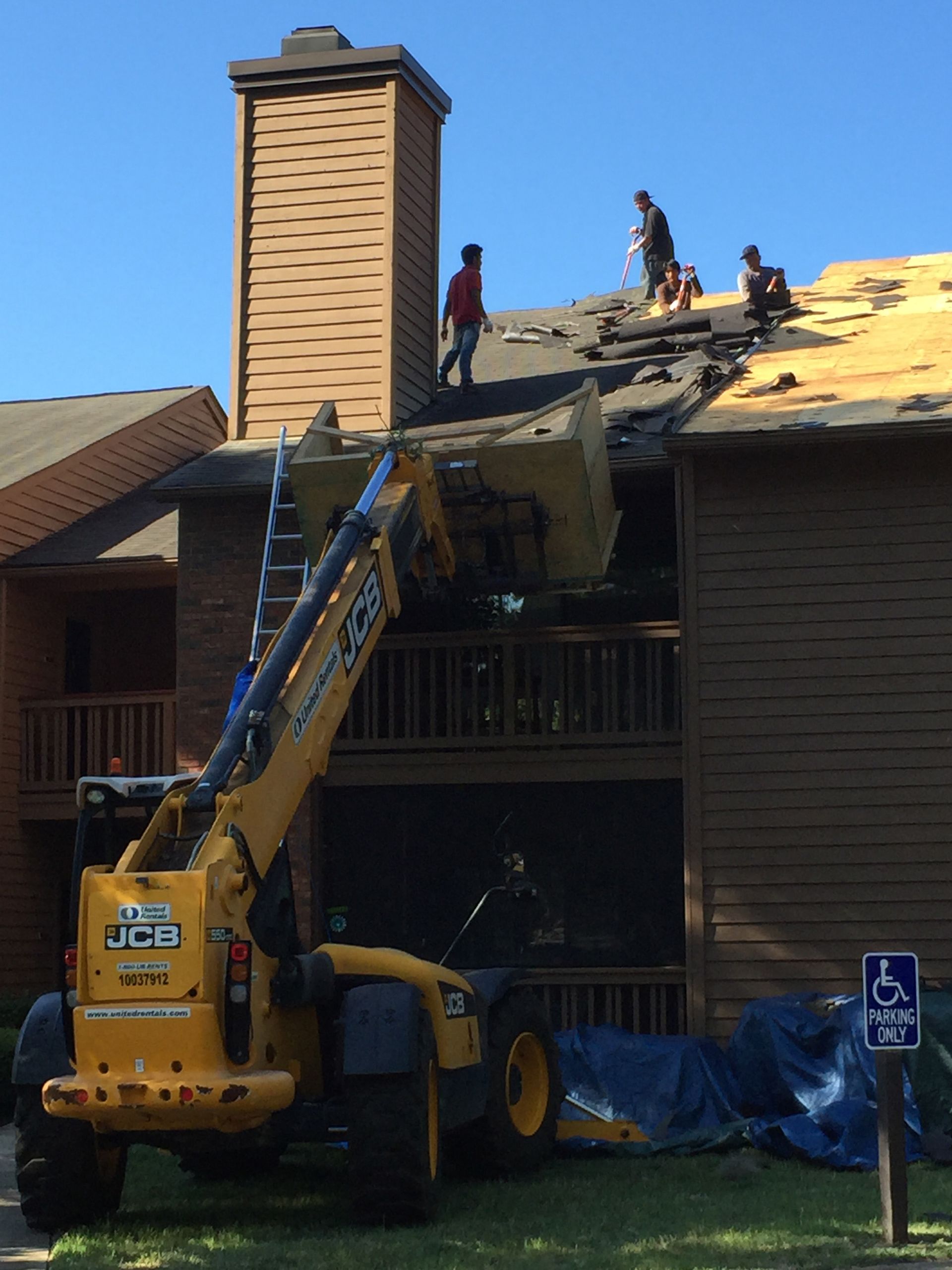 Roofers removing shingles from an apartment building roof using a yellow JCB telehandler.