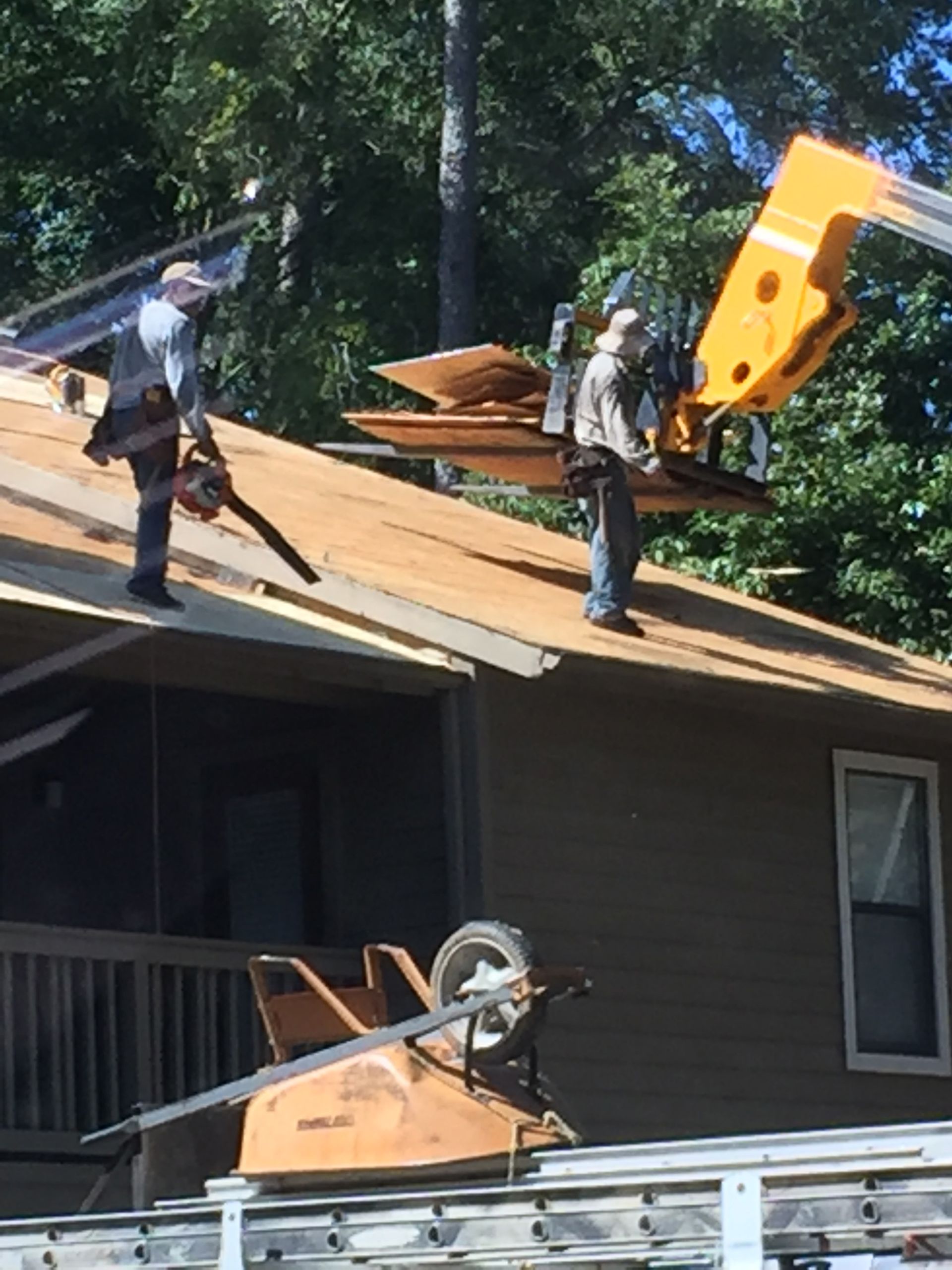 Two workers on a roof, one cutting with a chainsaw, another by a lifting device, replacing shingles.