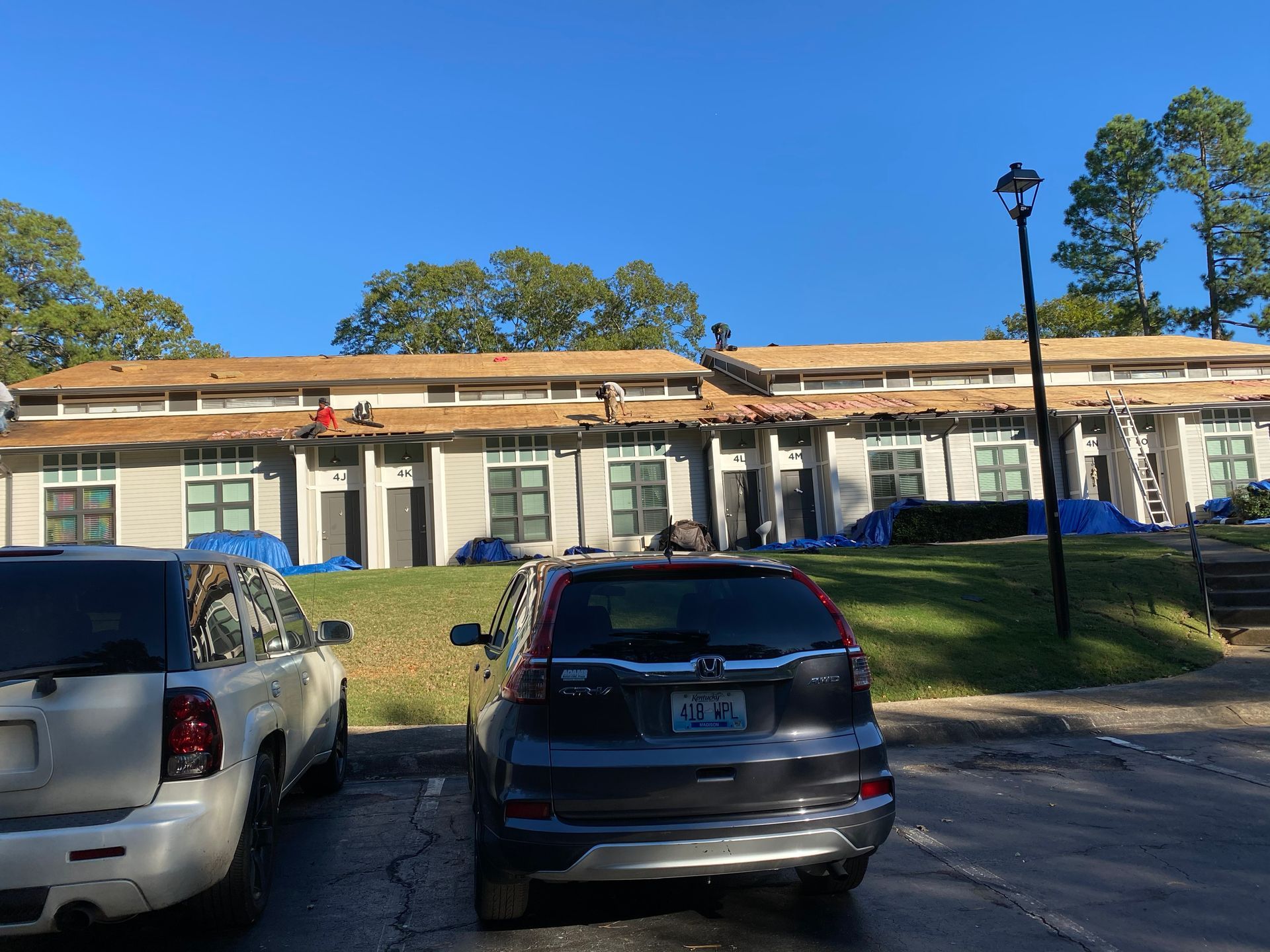 Apartment buildings with roof construction; workers on roofs, cars parked below. Blue sky.