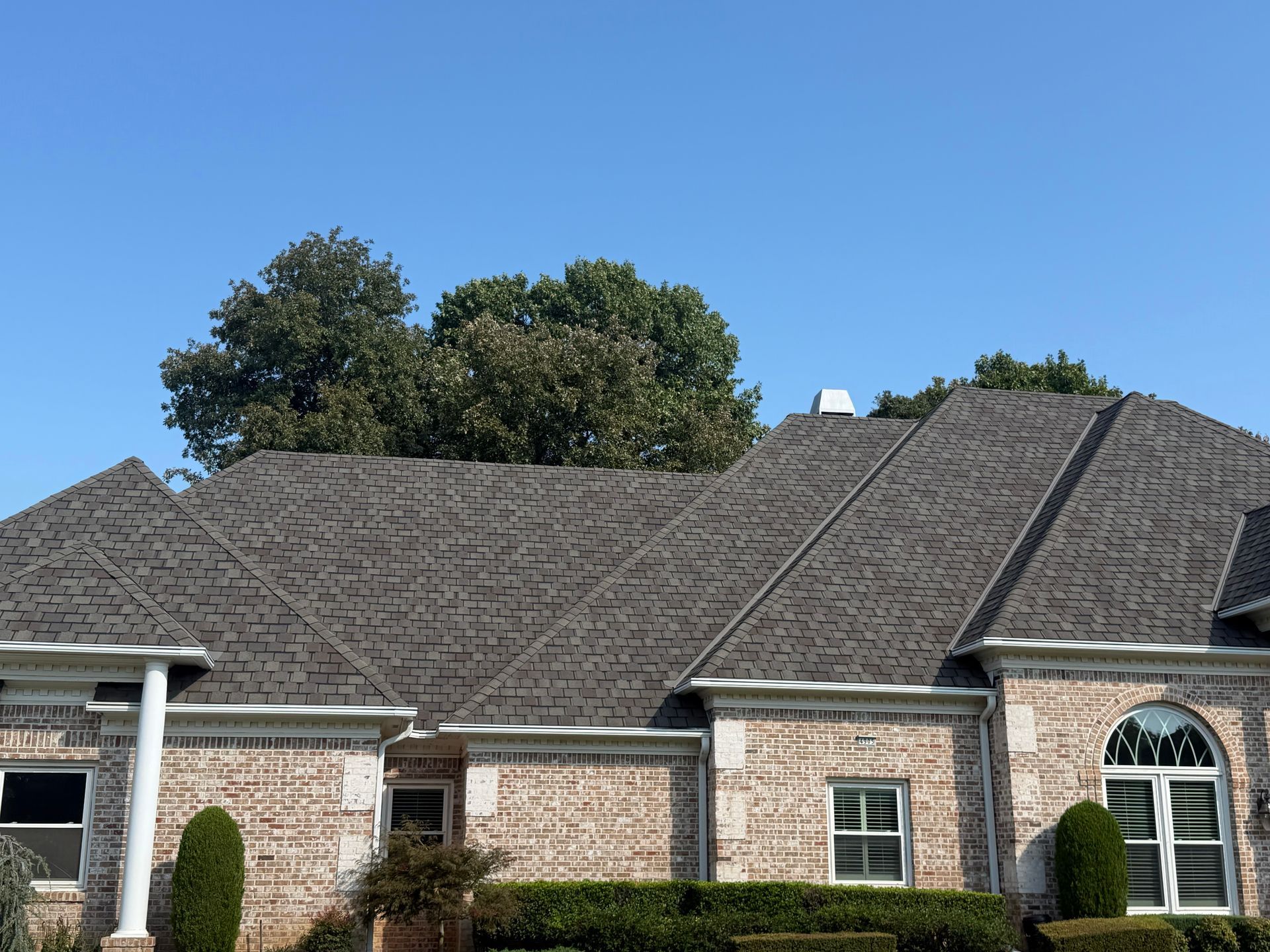 Brick house with dark gray shingle roof under a blue sky, some trees in the background.