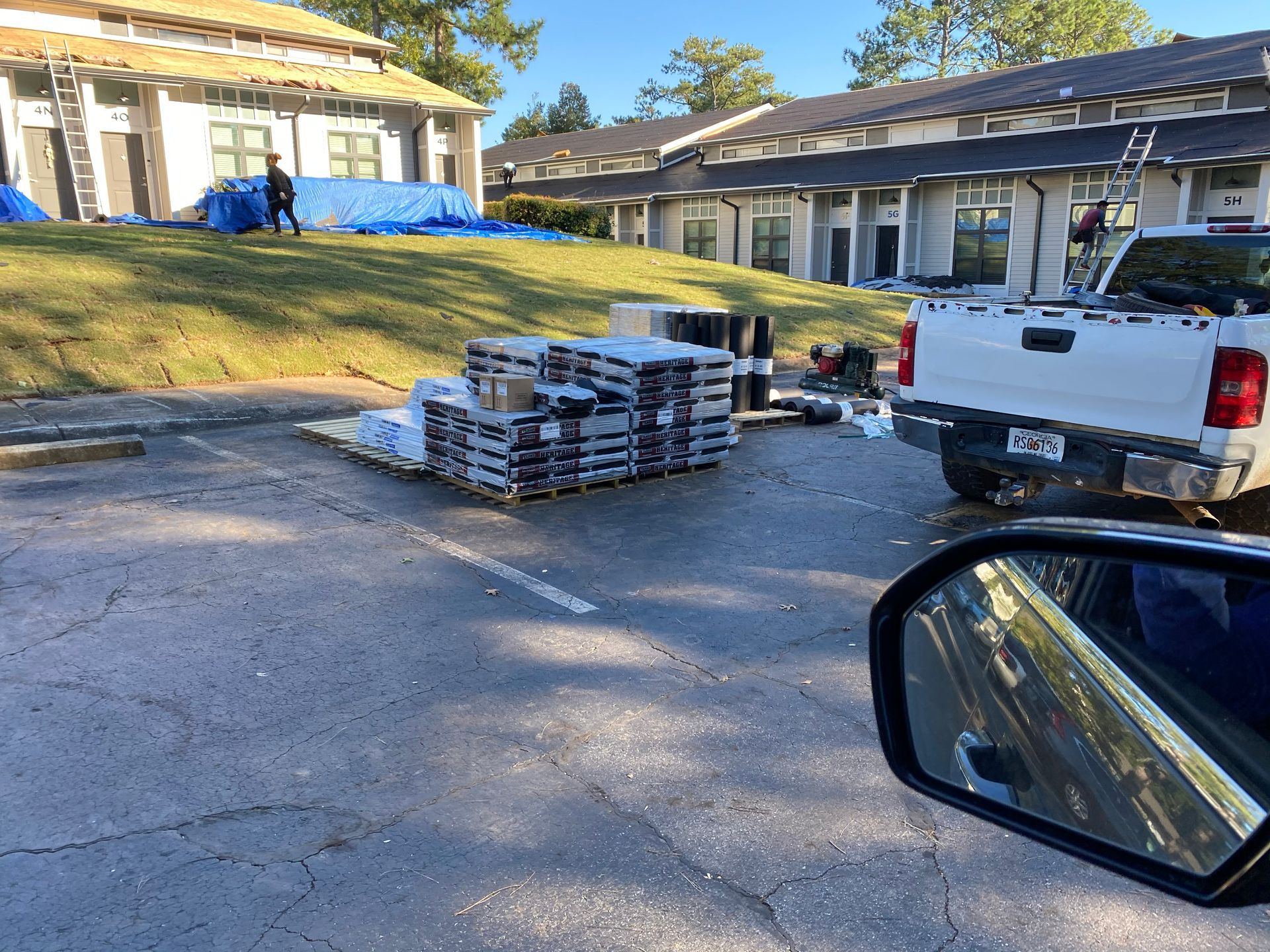 Roofing supplies on pallets in a parking lot next to buildings and a truck. Workers and tarps on the roof.