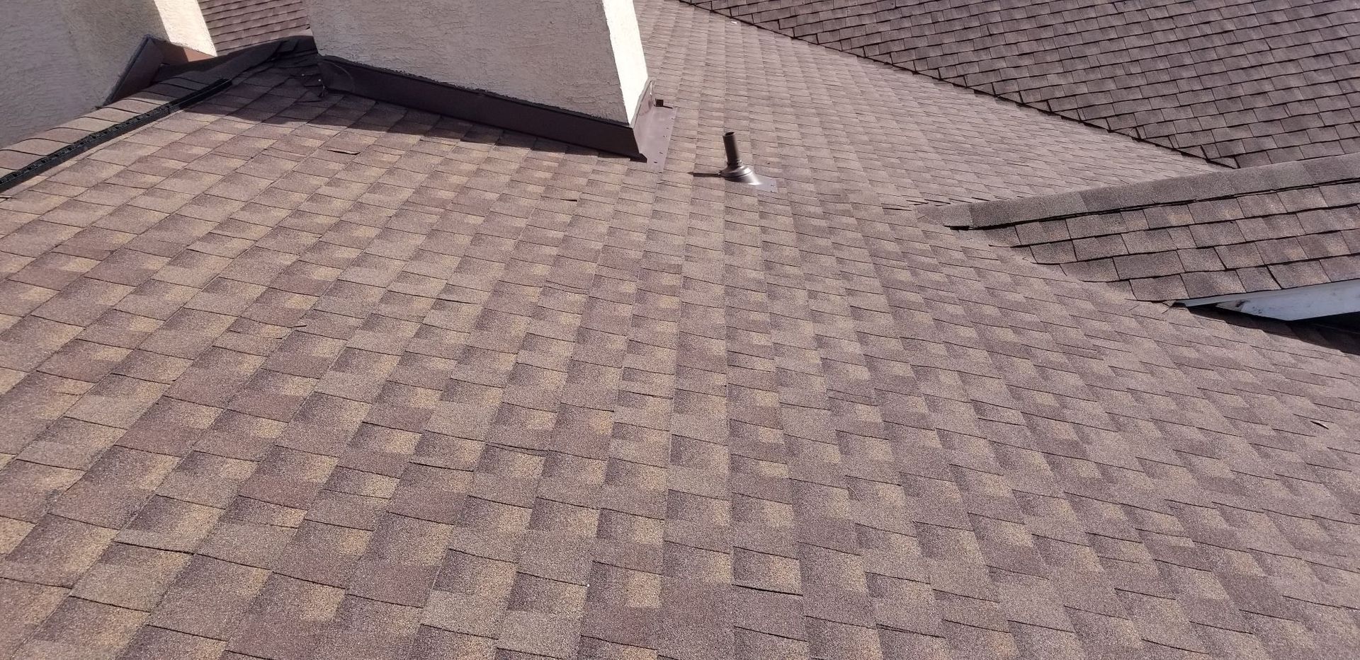 A brown shingle roof with a vent pipe. The roof has multiple angles and a light-colored wall is visible.