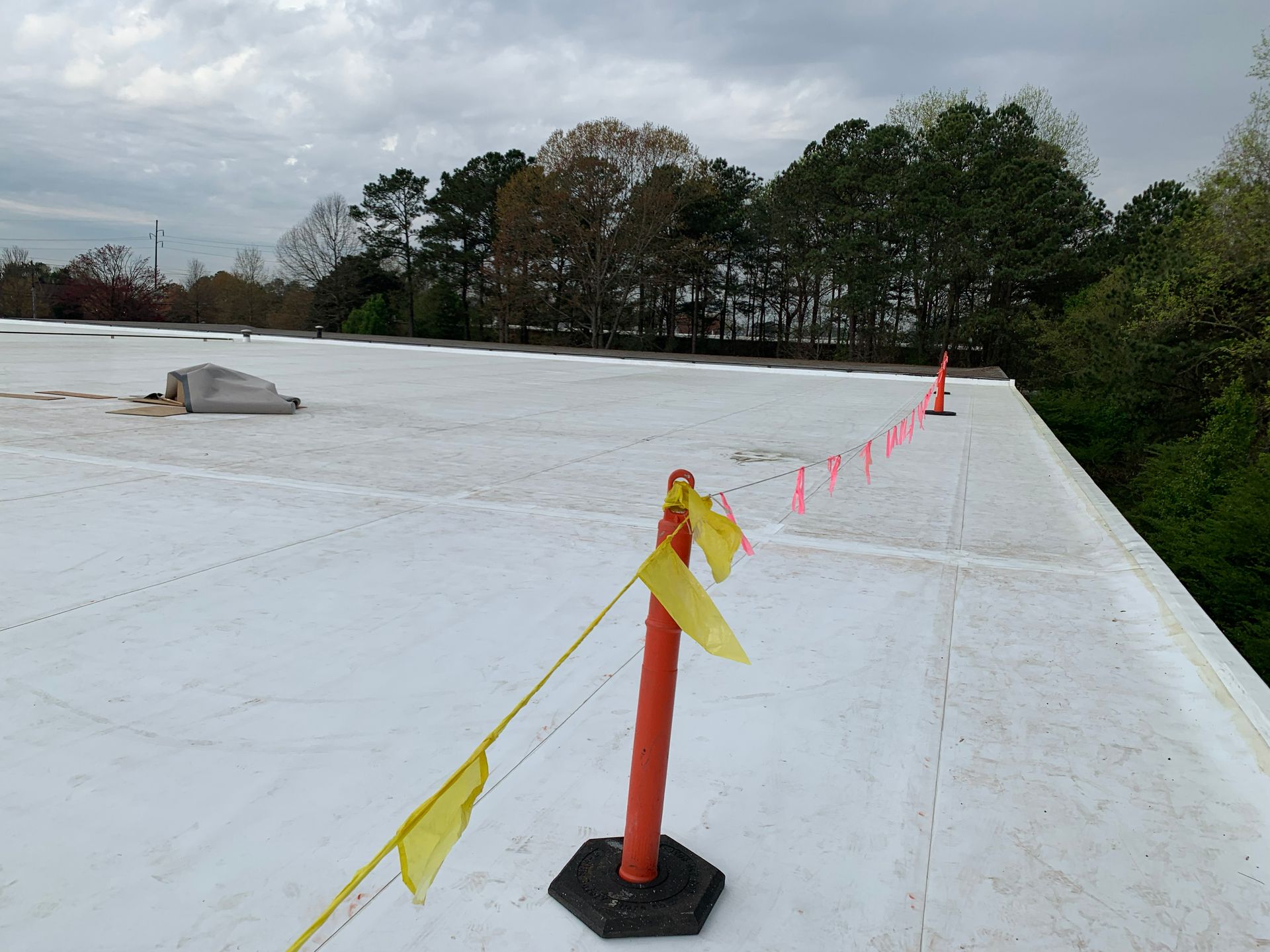 Flat roof with safety line, orange cones, and pink string. Trees and cloudy sky in the background.