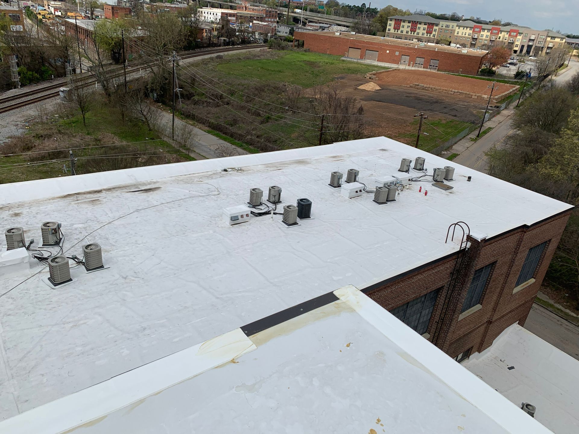 View of a flat, white commercial roof with HVAC units. Brick building, dirt lot, and railroad tracks in the background.