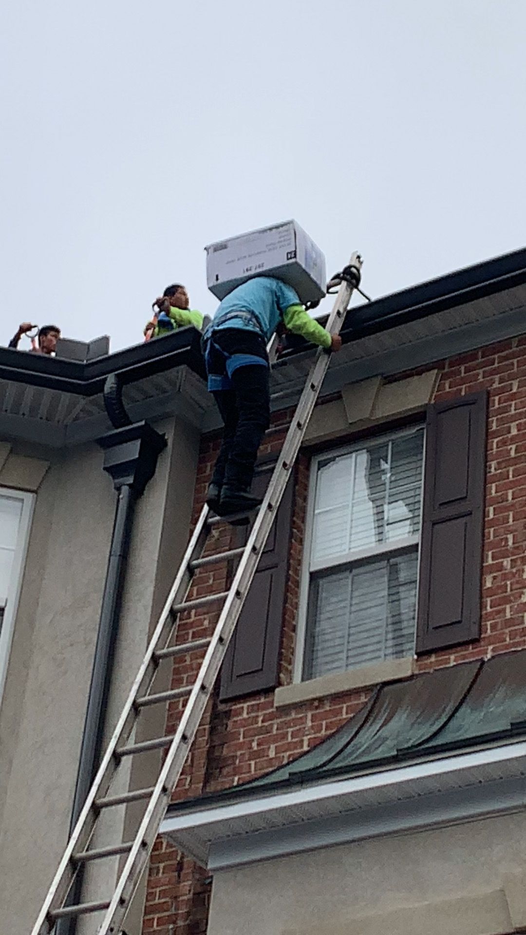 Man on ladder carries a box on his head. Two people on roof. Building with brick and gutter.