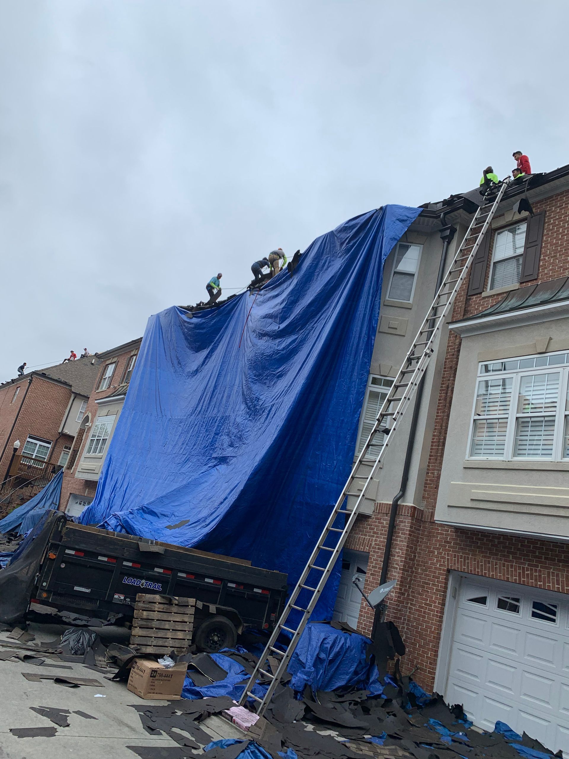 Workers covering a damaged roof with a large blue tarp. A ladder and debris are also present.