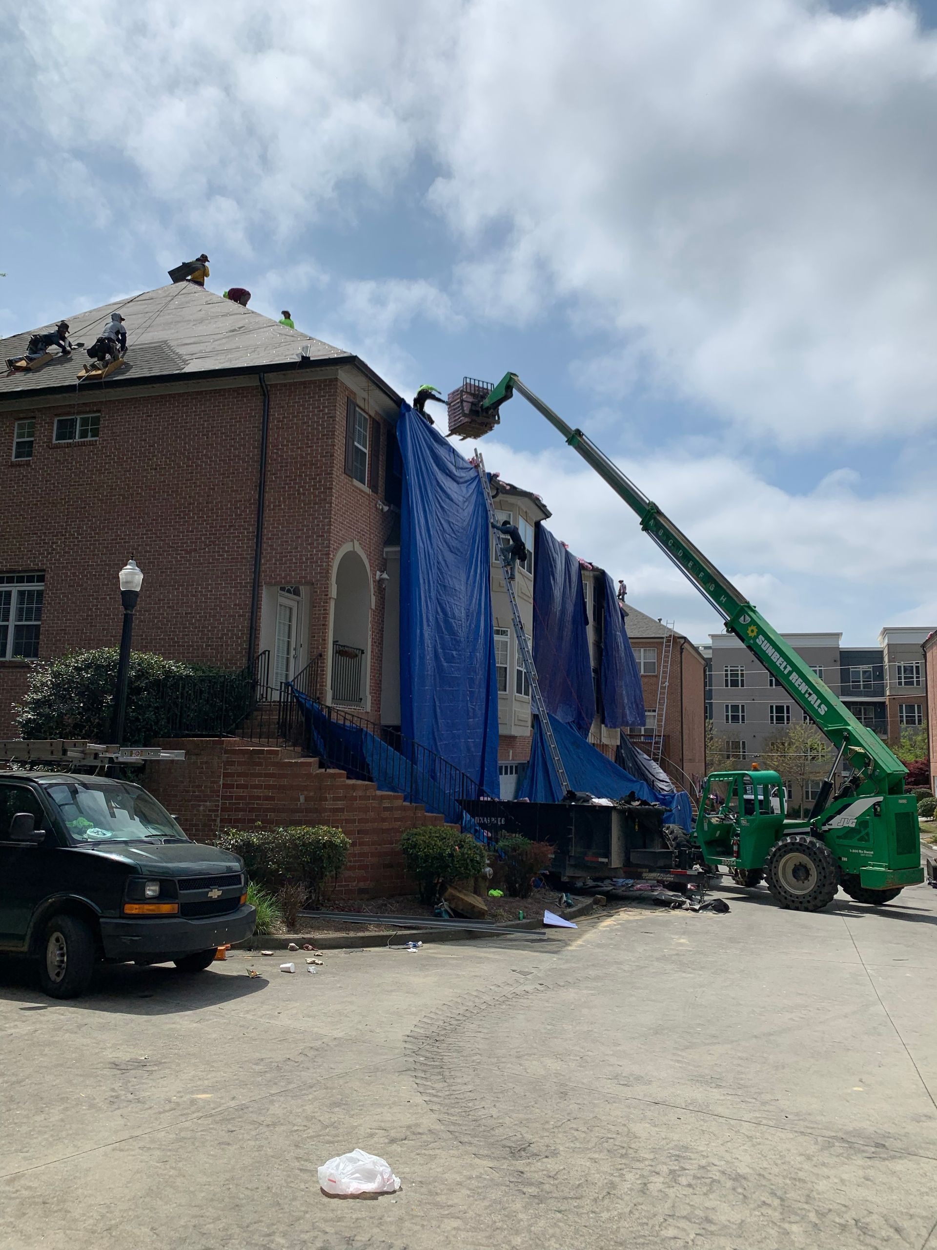 Damaged brick building with blue tarp being installed by a green lift. A black van is parked nearby.