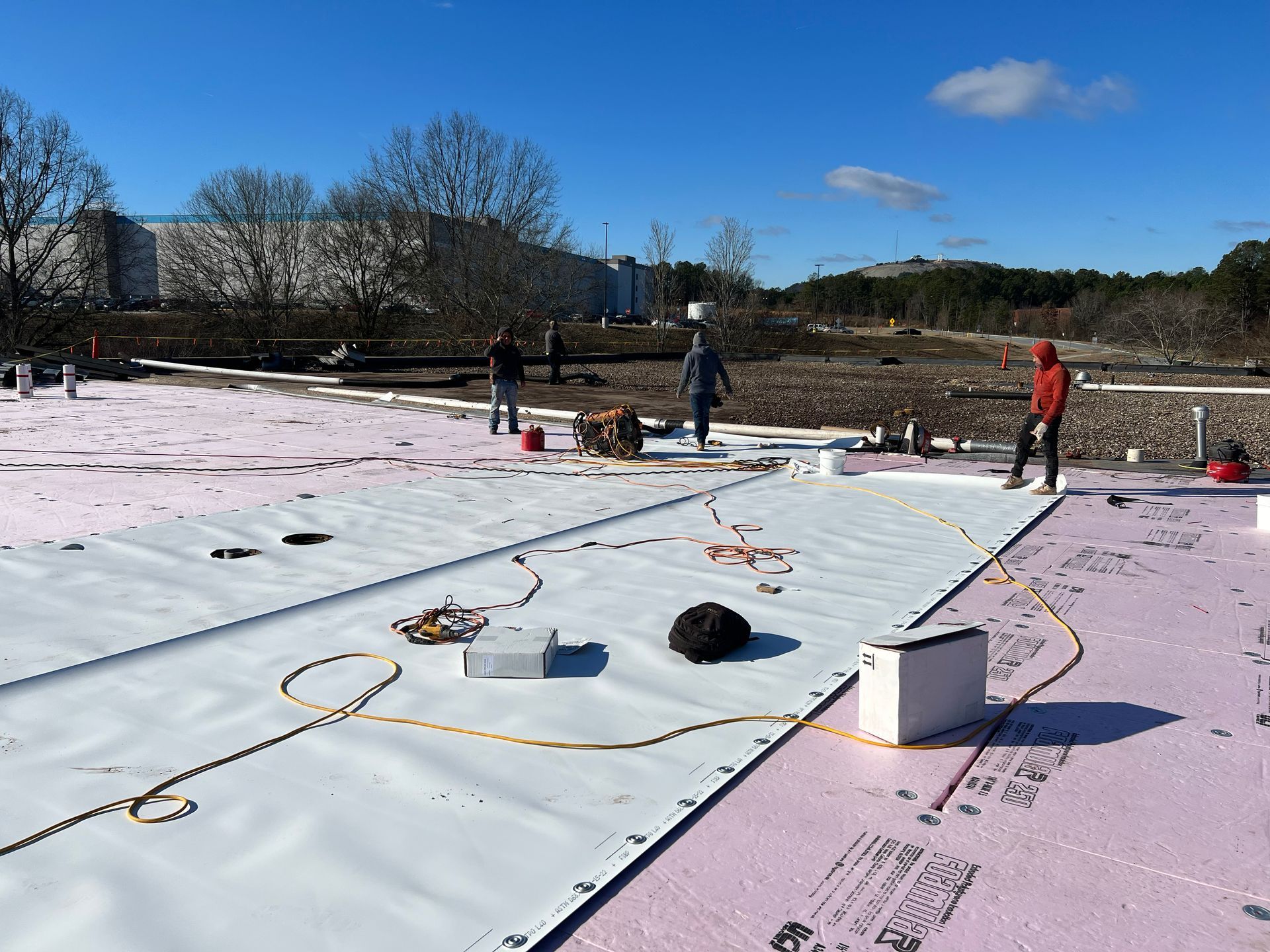 Workers on a flat roof installing white membrane, pink insulation, under a blue sky.