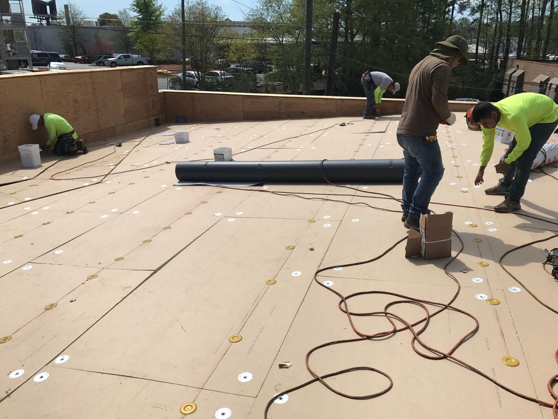Construction workers on a rooftop installing material; beige surface, multiple workers, power cords, sunny day.