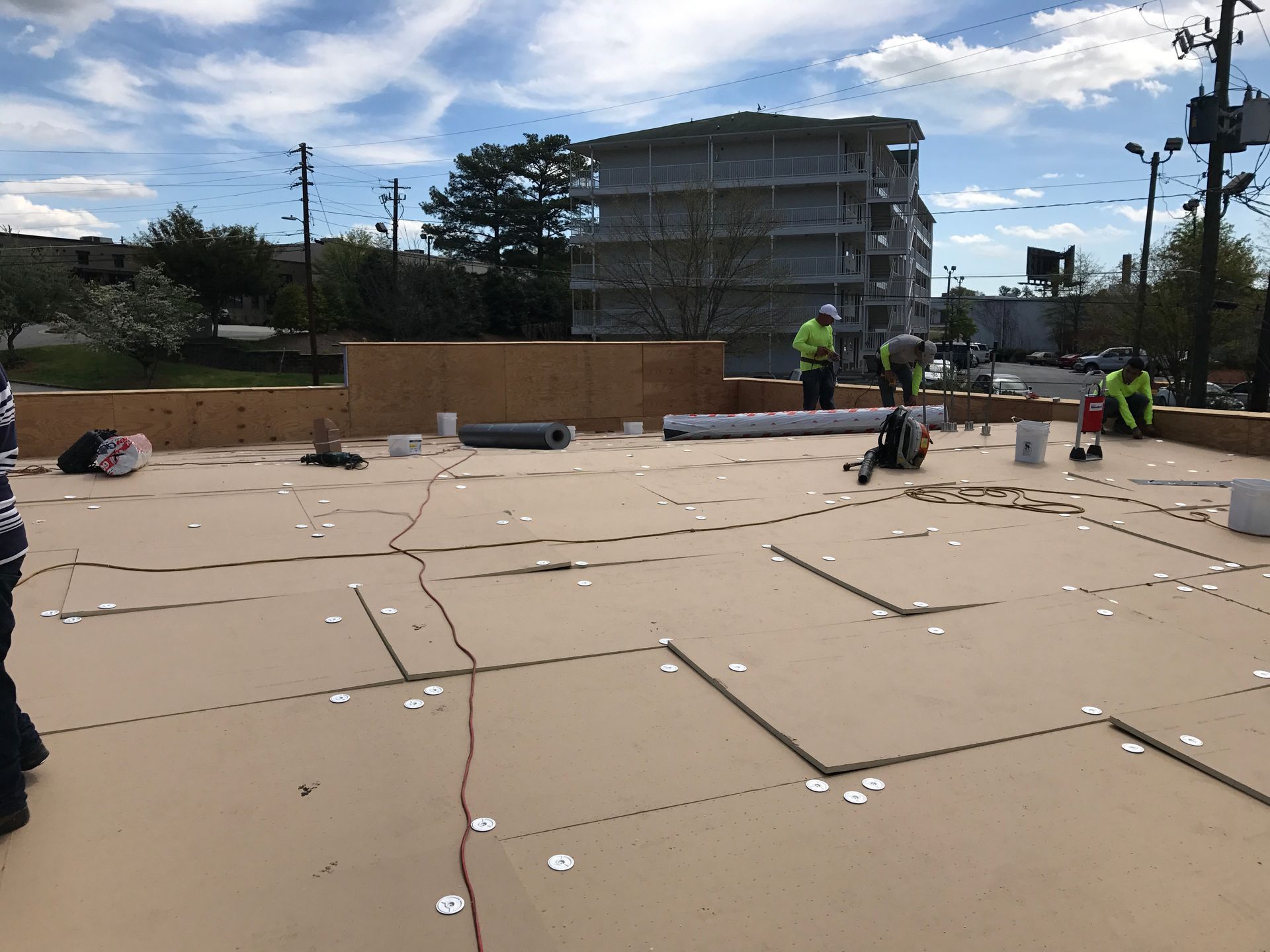 Construction workers on a rooftop, installing materials. Light-colored surface with many markings, surrounded by cityscape.